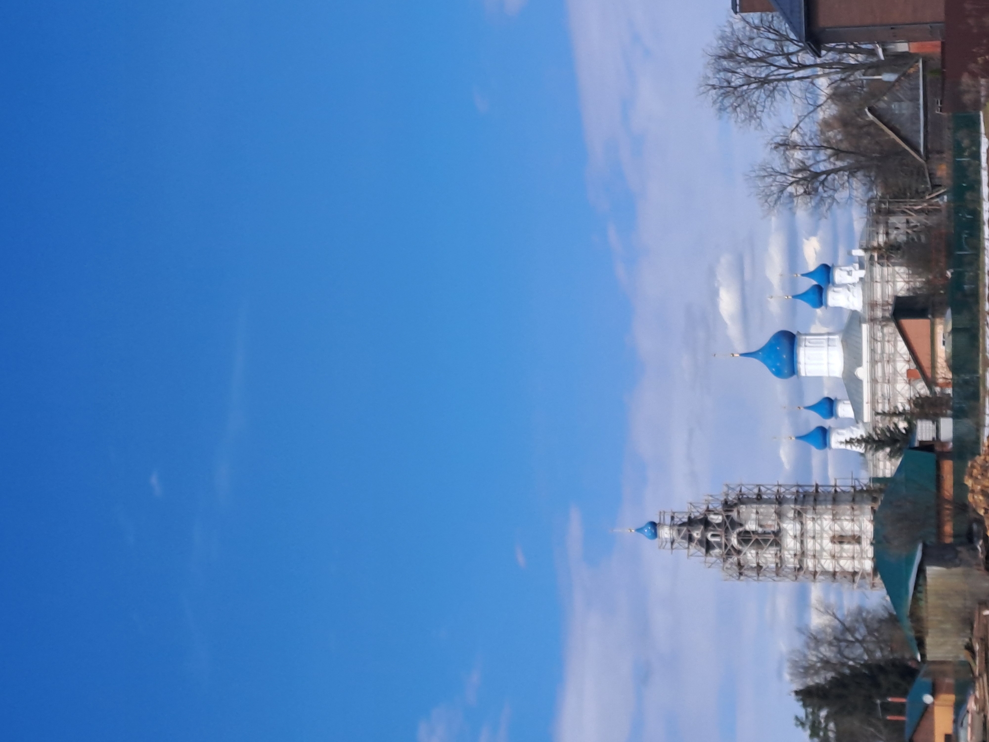 The image depicts a Russian Orthodox church with blue domes, possibly undergoing restoration, as it's surrounded by scaffolding. The church is located in Казанское, Russia. The sky is blue with wisps of clouds. Trees and buildings are visible in the foreground.