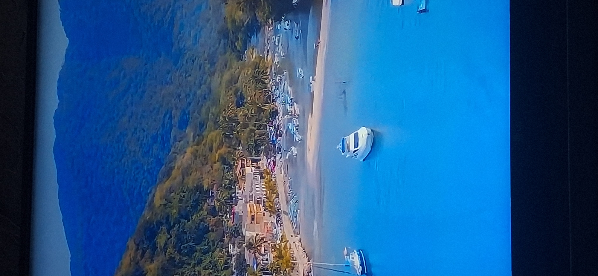 The image depicts a coastal town nestled at the base of a steep, densely vegetated mountain. A wide bay filled with calm, bright blue water dominates the foreground. Several boats are visible on the water, including a white motor yacht in the center and a blue and white sailboat to its left. Along the shoreline, a sandy beach is lined with buildings, appearing to be a mix of hotels and shops, interspersed with palm trees. Further out in the bay, a multitude of smaller boats are moored. The mountain rises sharply behind the town, its slopes covered in lush green foliage and rocky outcrops, fading into a hazy blue sky at the summit. There is no indication that this scene is located in Bila Tserkva, Ukraine, as it appears to be a tropical or subtropical coastal environment.
