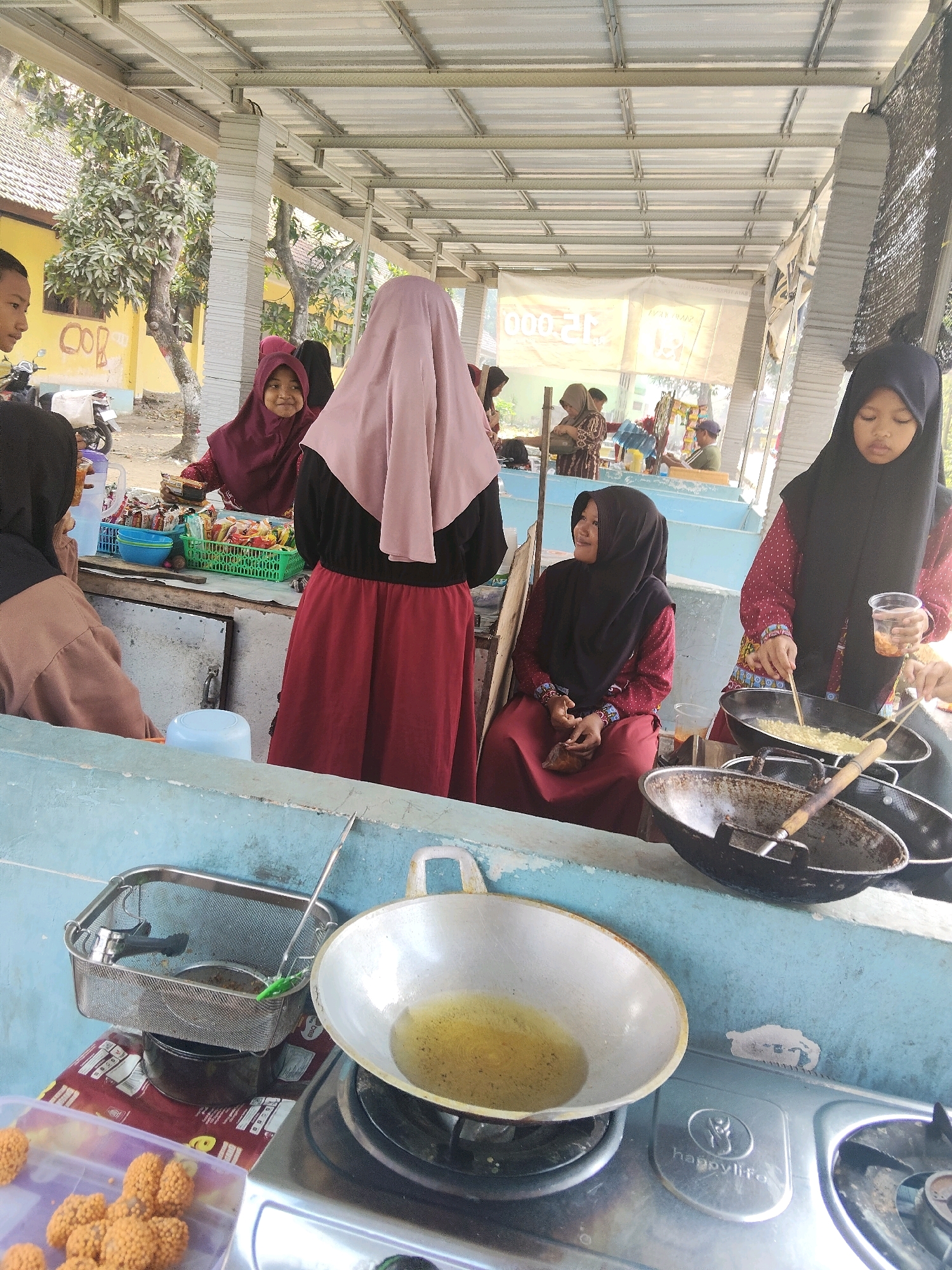 In Payungrejo, Indonesia, a group of young Indonesian girls in hijabs are gathered at an outdoor food stall. A young girl in a maroon hijab and dress is actively engaged in cooking, using chopsticks to stir food in a black wok placed on a gas stove. Beside her, another girl in a black hijab and red patterned top holds a plastic cup and watches the cooking. In the foreground, a light blue counter displays cooking utensils, including a deep frying basket filled with oil, and a large white wok with oil. To the left, a plastic container holds fried food items. In the background, more individuals are visible near a stall with baskets of snacks, and a yellow building and trees suggest a rural or suburban setting. The overall scene captures a moment of daily life at a bustling local food vendor.