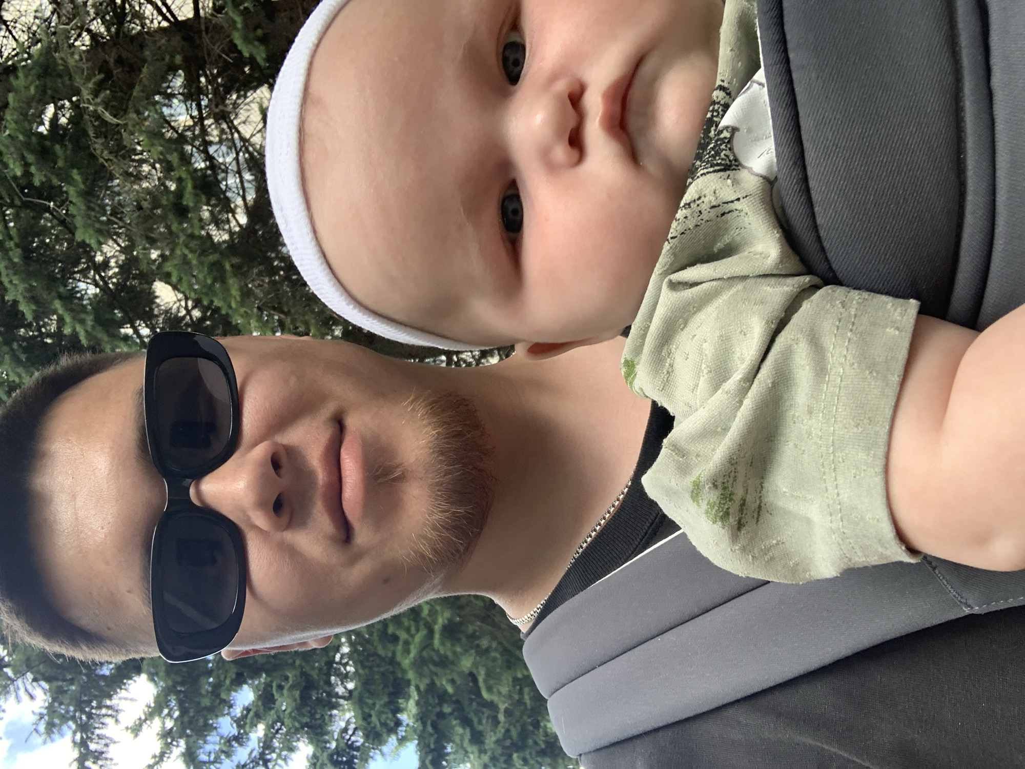 This is a close-up, selfie-style photo taken outdoors, likely during the daytime. The image features a man and a baby, possibly father and child, with a background of green trees and a cloudy sky, hinting at a temperate climate.
The man is wearing black sunglasses and has a light beard. He is in the foreground, positioned to the left of the frame. A baby, wearing a white beanie, is held in a carrier against the man's chest. The baby's face is visible, showing soft features and dark eyes. The baby is wearing a light green t-shirt with a textured pattern. The man is wearing a black t-shirt with a silver chain visible around his neck. The baby carrier is dark gray.
The overall mood appears casual and intimate, suggesting a moment of shared outdoor activity between the man and the baby. The lighting suggests it is daytime, with natural light illuminating the subjects. The foliage in the background indicates a natural, possibly park-like or wooded, setting. There is no visible text in the image.