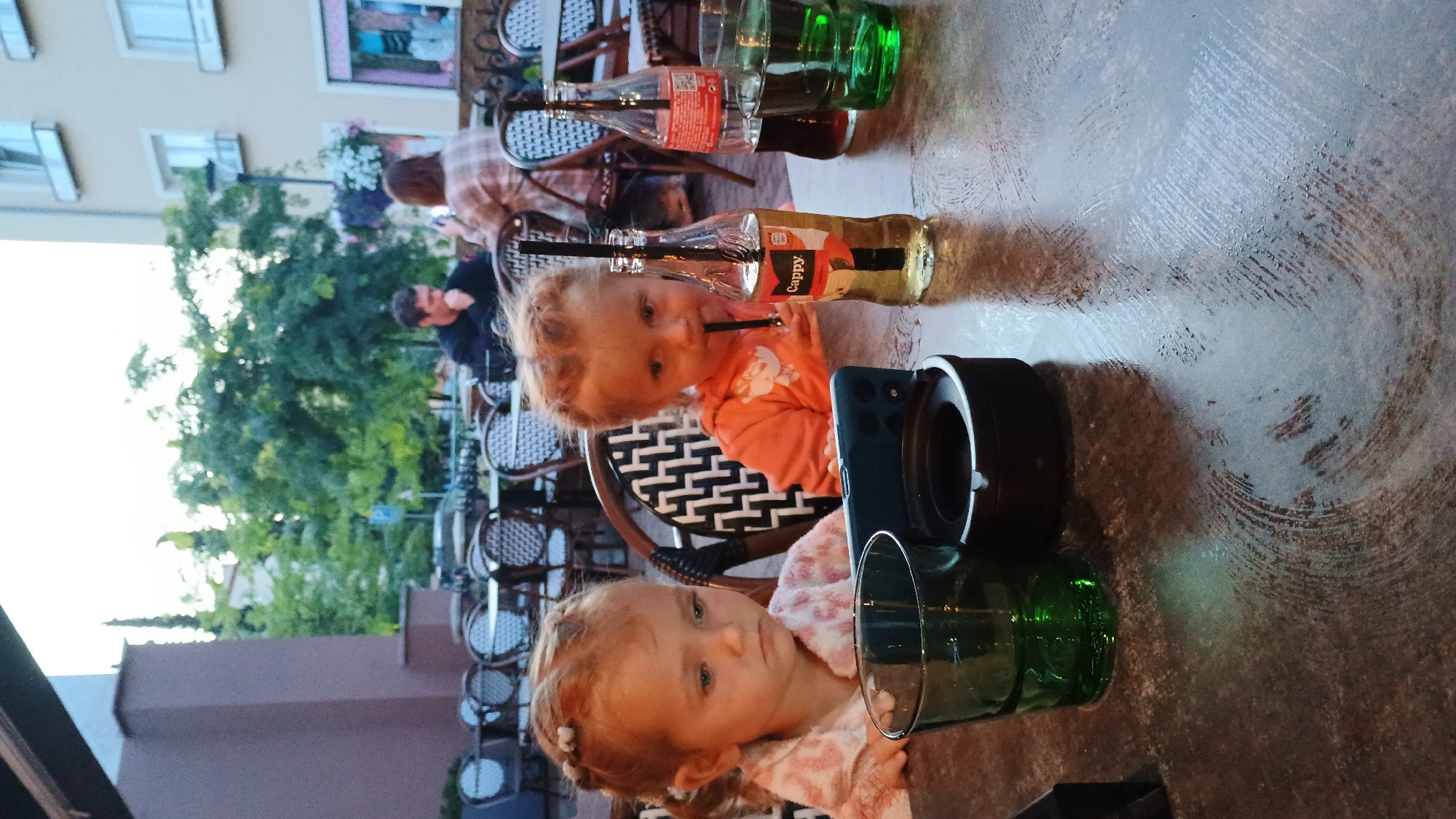 In an outdoor seating area of a restaurant in Sokolov, Czechia, two young girls are seated at a table. The girl on the left, with blonde hair in pigtails, wears a pink and white patterned shirt and gazes downwards, seemingly engrossed by a smartphone propped up on the table in front of her. Beside her, a green glass holds a beverage. To her right, another young girl, also with blonde hair in pigtails, wears an orange hooded sweatshirt. She is actively drinking from a bottle of Cappy juice with a straw, her attention also directed towards the phone. On the table, near her, are the Cappy bottle, another bottle of dark liquid (likely cola), and a green glass containing a dark beverage. An ashtray is positioned between the two girls. In the background, other patrons are visible seated at tables, along with trees and buildings, indicating an urban environment. The lighting suggests it is late afternoon or early evening.