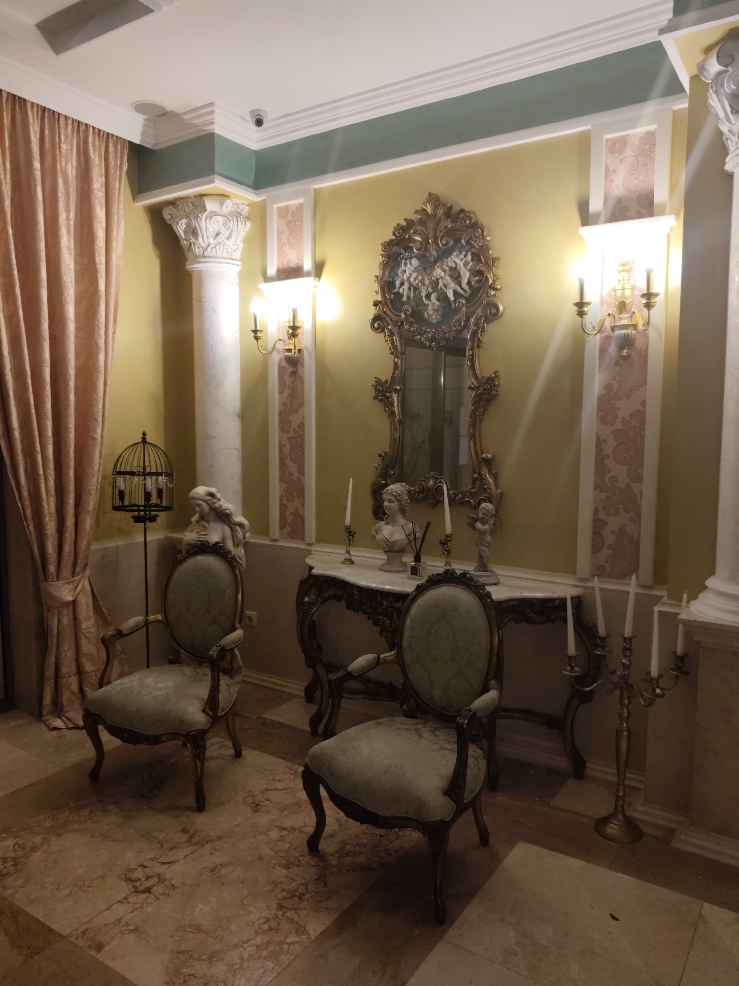 The image shows an interior scene, likely within a building in the historic city of Nesebar, Bulgaria. The room is decorated in a classical style, with ornate furnishings and architectural details.
Dominating the background is a large, gilded mirror with intricate carvings, reflecting the ambient light. Flanking the mirror on the walls are two decorative sconces, each with two candle-like lights. The walls are painted a warm yellow, with accents of a lighter patterned wallpaper. Two large, white Corinthian columns stand on either side of the room, adding to the grand aesthetic.
In the foreground, two upholstered armchairs with pale green patterned fabric and dark wood frames are positioned. To the left of the chairs, a decorative birdcage with three visible candles inside sits on a stand. Between the chairs and the mirror is a marble-topped console table, also ornately carved. On the table are several decorative items, including two busts (one of a woman and one of a cherub), a tall candle in a holder, and a diffuser with reeds.
To the right of the console table, a tall, multi-branched candelabra with four unlit white candles stands on the floor. The floor is tiled with a marble pattern in shades of beige and pink. A light pink curtain is visible on the far left of the image, partially obscuring the wall.
The lighting appears to be artificial, casting a warm glow throughout the room and creating soft shadows. There are no people visible in the image. The overall impression is one of opulence and historical charm.