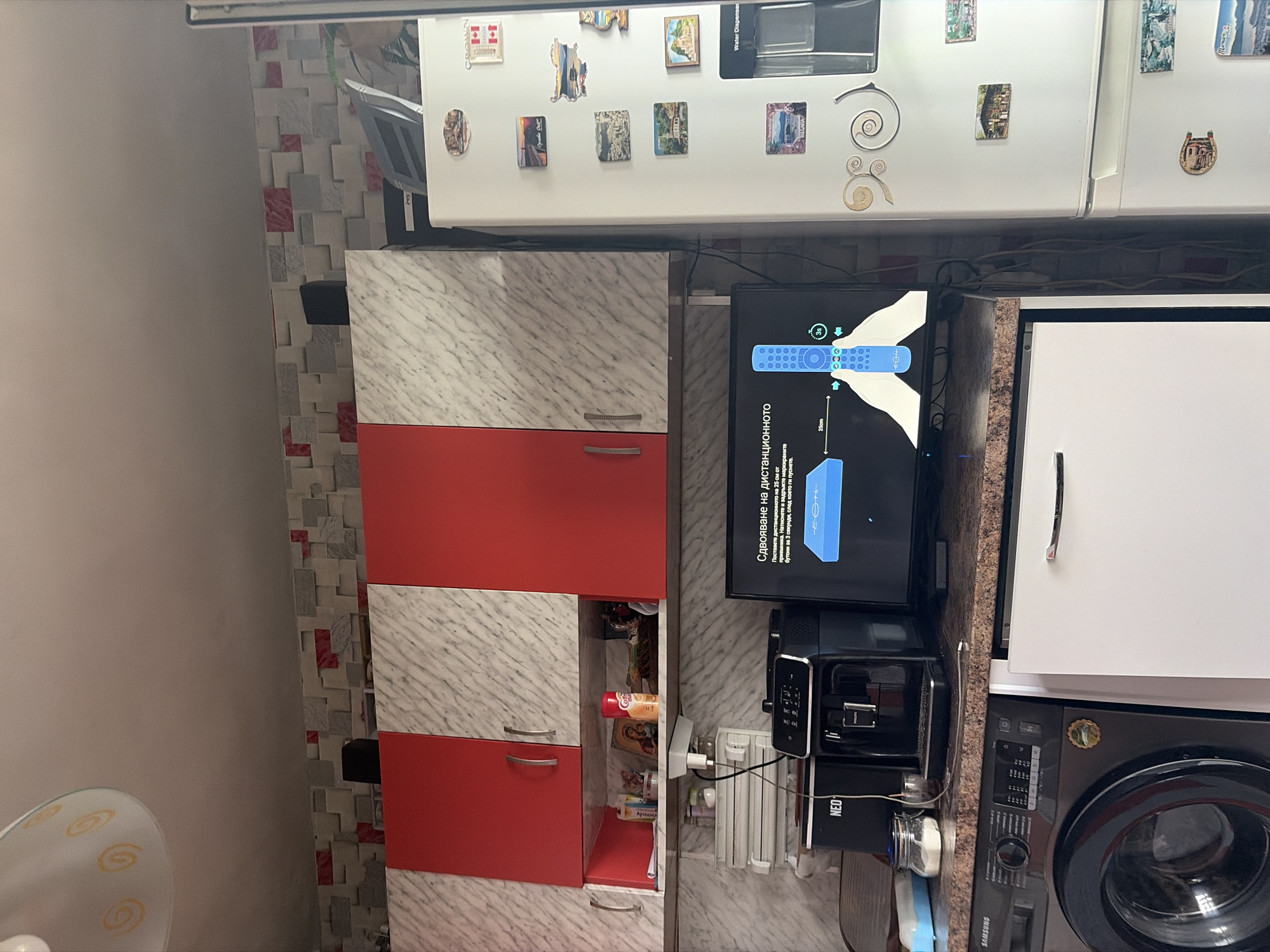 A kitchen scene. A washing machine is situated under a kitchen cabinet, next to a countertop supporting an oven and a television that displays connection instructions in Bulgarian. Cabinets of red and marble patterns are adjacent to the appliances. The refrigerator above is decorated with magnets, adding a personal touch to the space. The location appears to be Varna, Bulgaria, based on a "Varna" magnet on the refrigerator.