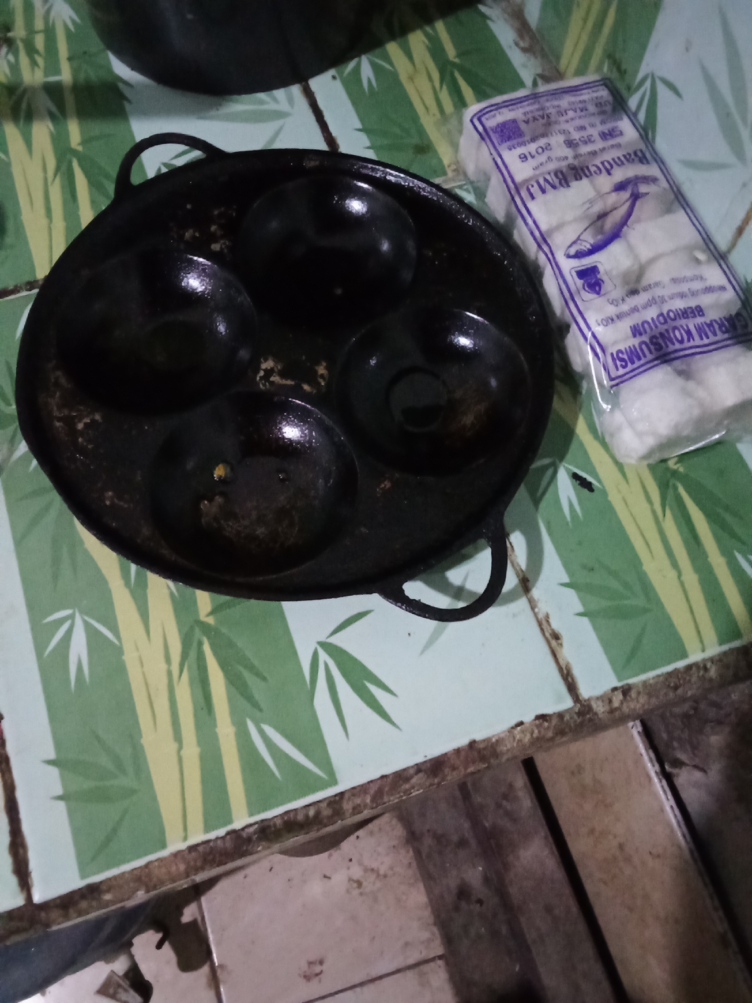 A black, cast-iron pan with four circular indentations is placed on a green and yellow tiled surface decorated with a bamboo leaf pattern. Next to the pan is a clear plastic package containing white, rectangular food items, possibly snacks or crackers. The packaging has purple text printed on it, including the words "Bandeng BMJ" and "GARAM KONSUMSI". The food items inside appear to be uniformly shaped and white in color. The tiled surface appears slightly dirty in some areas. The background shows a dark object, possibly another cooking utensil. The image is taken from a top-down perspective. The scene is likely indoors, possibly in a kitchen, as suggested by the cooking equipment. Without further context, the exact location in Pemalang, Indonesia, cannot be determined from the visual information alone.