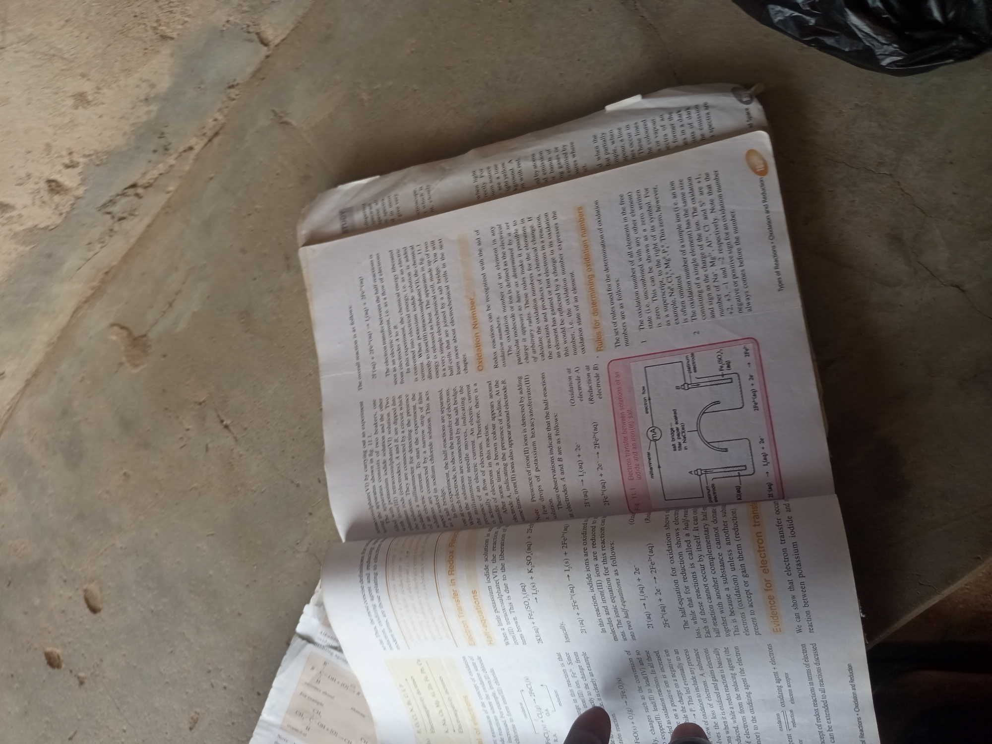 The image shows an open book displaying chemical equations related to oxidation and reduction. The book is lying on a rough, light-colored surface, possibly concrete or textured flooring. The lighting suggests an indoor environment. There are no people or explicit signs of the city of Jalingo, Nigeria. The focus is on the printed material and the immediate surface it rests upon.
