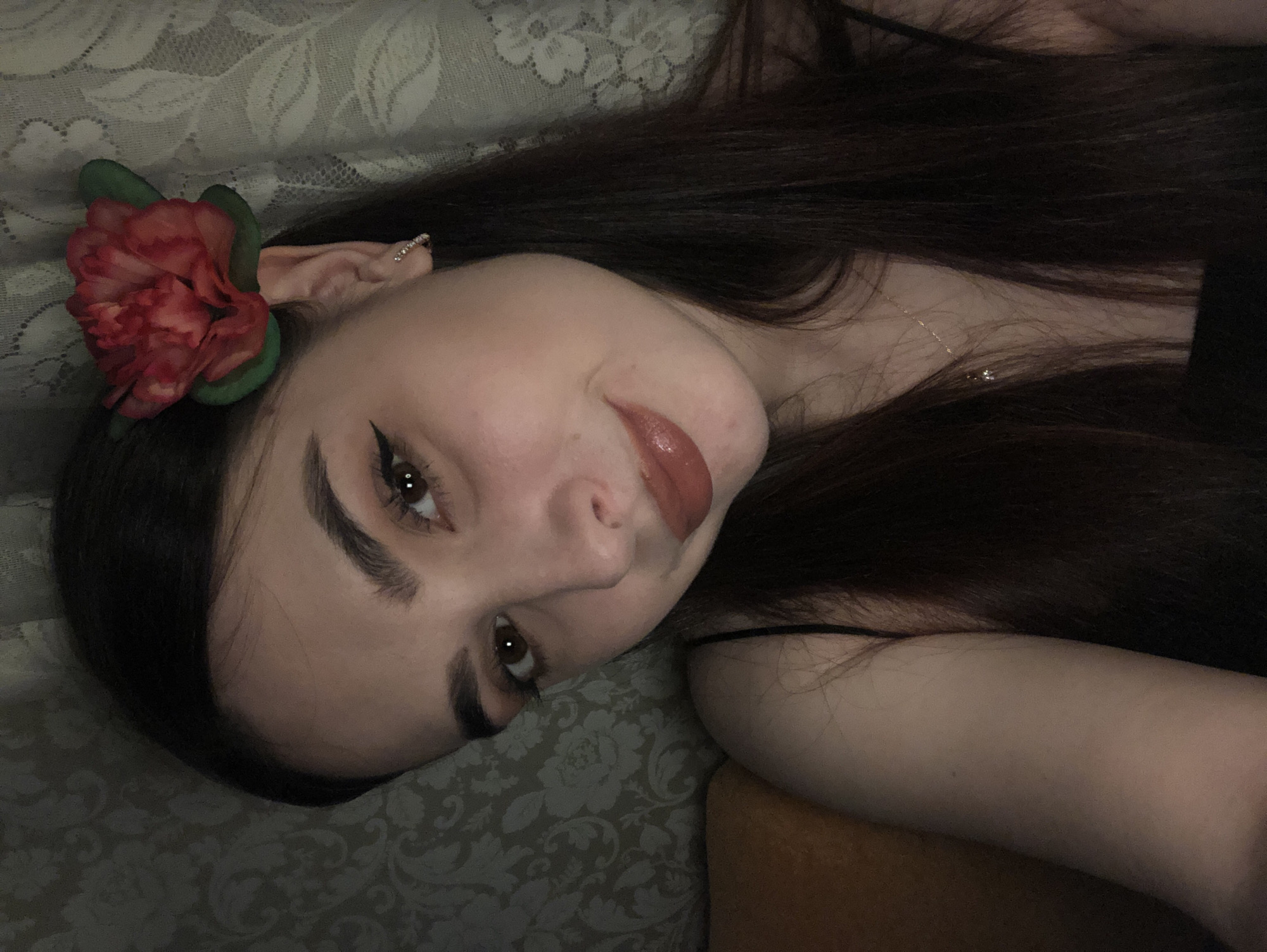 A woman with fair skin and long, dark brown hair is lying down, her head tilted towards the left. She has a red flower attached near her left ear, which appears to be slightly pointed and adorned with a small earring. She is wearing eyeliner, and her lips are coated in glossy, dark lipstick. A thin necklace is visible on her neck. She is lying on a patterned, gray surface, and a portion of her right arm rests on an orange-colored object. The image does not contain information about the city of Noviy, Russia.