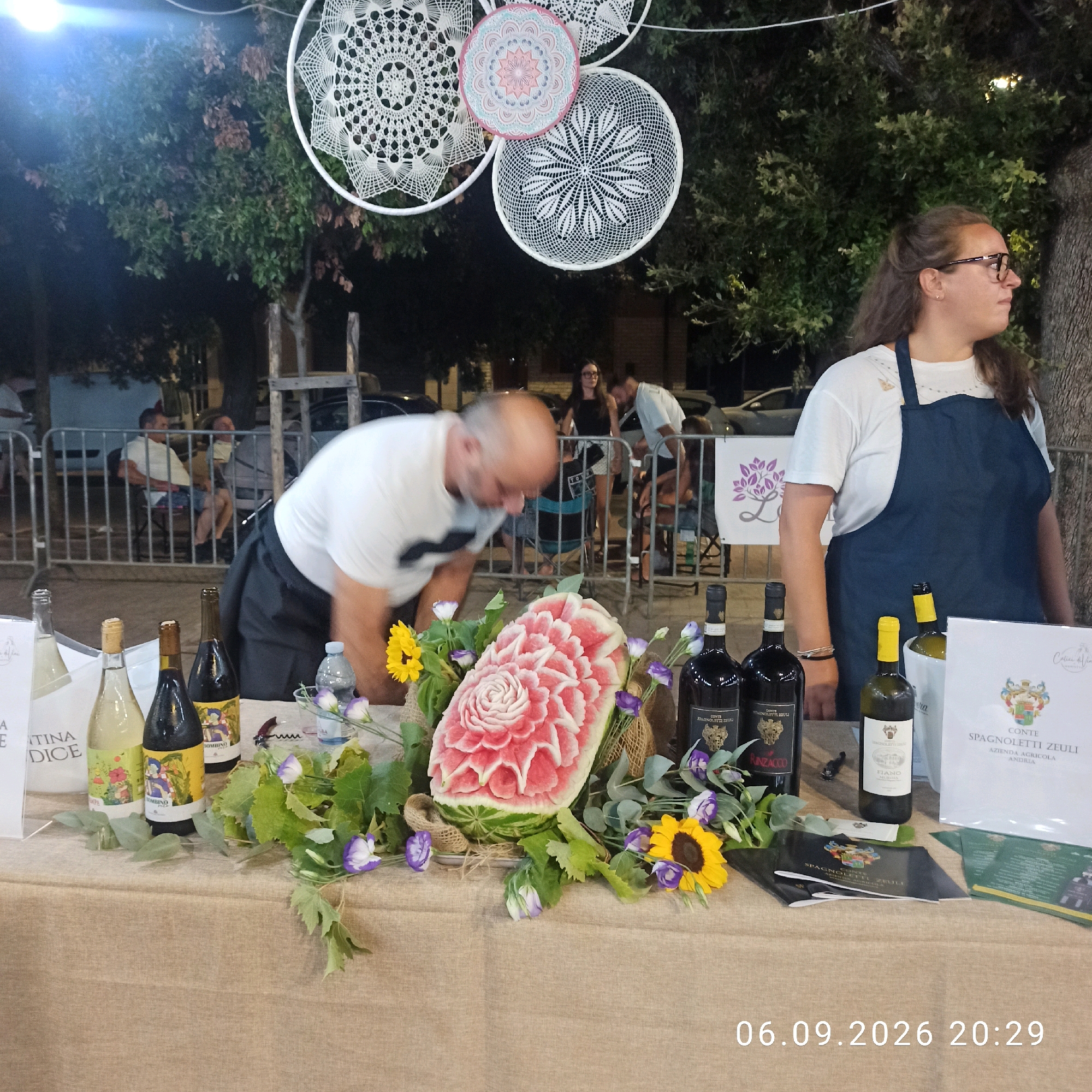 In Bitonto, Italy, a table is set with bottles of wine, a skillfully carved watermelon resembling a flower, and decorative floral elements. A man in a white t-shirt and shorts is leaning over the table, potentially arranging the display or attending to the carved watermelon. Behind the table, a woman wearing glasses and a dark apron stands, suggesting she might be a vendor or staff member. In the background, other individuals are visible, some seated behind a metal barrier, and trees form a natural backdrop. The scene appears to be an outdoor event or market, possibly related to a wine tasting or local festival, with decorative circular hangings above the display. The timestamp "06.09.2026 20:29" is superimposed on the lower right portion of the image.
