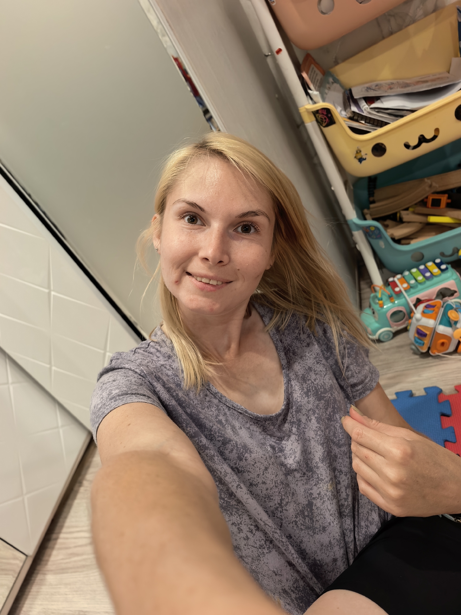A woman with blonde hair and fair skin is captured in a selfie, smiling at the camera. She is wearing a grey t-shirt with a speckled pattern. Her right arm is extended towards the camera, obscuring part of her body and the immediate surroundings.
In the background, a colorful storage unit filled with various items, including papers and toys, is visible. Among the toys are what appear to be wooden train tracks and a xylophone-like toy with colorful keys. The floor is wooden, and there are some interlocking foam play mats in red and blue. To the left of the woman, a wall with a tiled pattern is present.
The lighting suggests it could be daytime, possibly indoors given the presence of toys and storage units. The woman's expression is pleasant, indicating a casual and likely at-home setting. There is no visible text in the image.