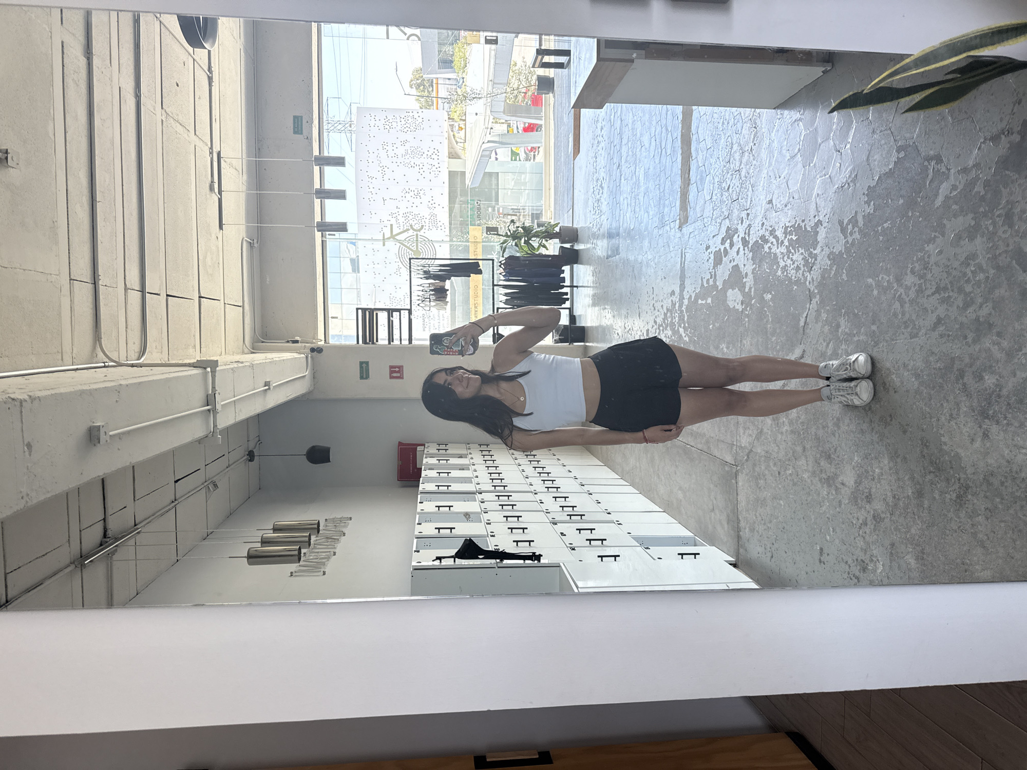 A young woman is captured in a full-body mirror selfie, standing casually inside what appears to be a modern studio or commercial space. She has long, dark hair and is dressed in a white sleeveless crop top, black shorts, and white sneakers, holding a smartphone to take the picture. A faint, relaxed smile is visible on her face.
The interior features a concrete floor with a textured, somewhat distressed appearance, and smooth concrete walls with exposed piping and electrical conduits. A long bank of white lockers with black handles lines the left side of the room. Modern, cylindrical metallic pendant lights hang from the ceiling, along with one darker, bell-shaped fixture.
Behind the woman, large windows offer a clear view of a bright, sunny street scene. Outside, urban buildings, including a distinctive white facade with numerous small perforations, are visible, along with parts of vehicles and trees. The word "STUDIO" is clearly reflected on the window, repeated and possibly part of a business name like "APEX STUDIO", suggesting the nature of the establishment. Other faint text, possibly "ROBIO", can also be seen. A large potted plant with variegated leaves is positioned to the right. The bright daylight indicates it is daytime with clear weather. The overall environment conveys a contemporary, functional, and possibly industrial aesthetic, characteristic of urban settings such as Puebla, Mexico.