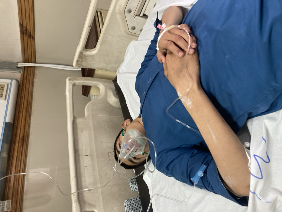 The image depicts a male individual, appearing to be a young adult or adolescent, lying supine on a hospital bed in a clinical environment within Quetta City Tehsil, Pakistan. The individual is wearing a dark blue long-sleeved garment. An oxygen mask is secured over his nose and mouth, connected via clear tubing extending upwards and to the left. An intravenous (IV) cannula with pink components is inserted into the dorsal aspect of his left hand, with clear tubing running from it. His hands are clasped together over his midsection. White residue, consistent with medical tape or disinfectant, is visible on both forearms. The bed features white bedding and a light-colored, possibly plastic or metal, frame showing some wear. The wall behind the bed is light-colored, with a wooden trim and a wall-mounted utility panel containing electrical outlets. White conduits are affixed to the wall next to the bed. A portion of a brown container is visible beside the bed on the right.