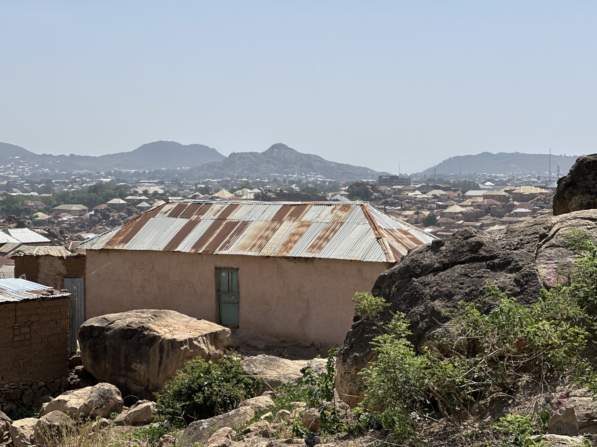 The image captures a scene in Jos, Nigeria, characterized by a landscape of rocky terrain interspersed with modest buildings. In the foreground, a small building with a corrugated metal roof and a light-colored exterior with a green door is prominently featured. The building is surrounded by large rocks and sparse vegetation. The background reveals a dense urban sprawl with numerous houses, gradually ascending towards a range of mountains. The overall atmosphere is hazy, suggesting a warm, dry climate. No people are visible.