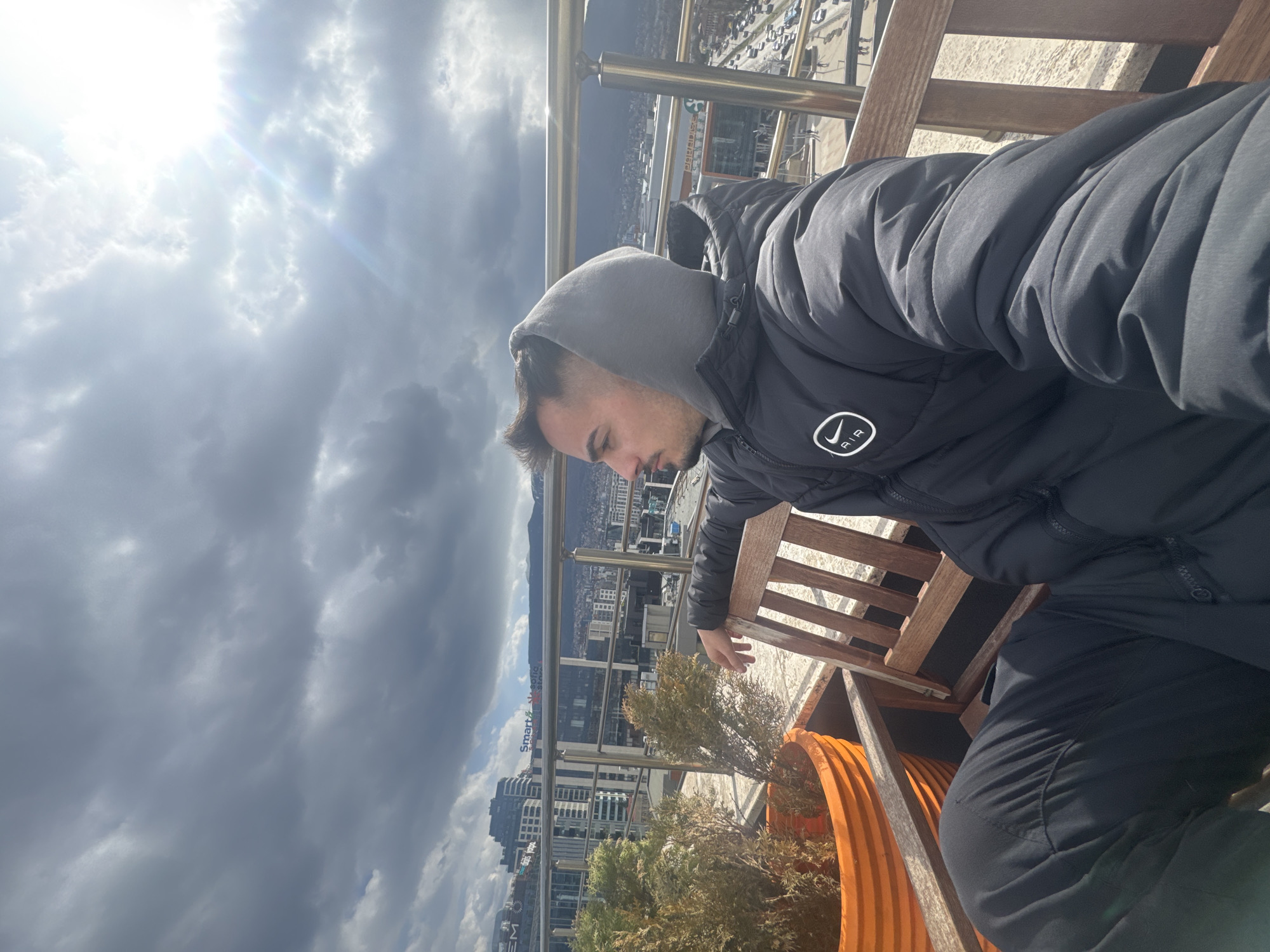 A man is seated on a wooden bench, gazing down at the city of Sofia, Bulgaria, from an elevated vantage point. He wears a grey hooded sweatshirt underneath a black puffy jacket with a circular logo containing an oval shape and the word "AIR". He is also wearing black pants. The foreground features an orange planter with a small bush. A metal railing separates the bench from the city view. In the background, Sofia is visible, with various buildings, mountains, and cloudy sky. The sky is partially covered with dark, heavy clouds with the sun peeking through, creating a contrast in light.