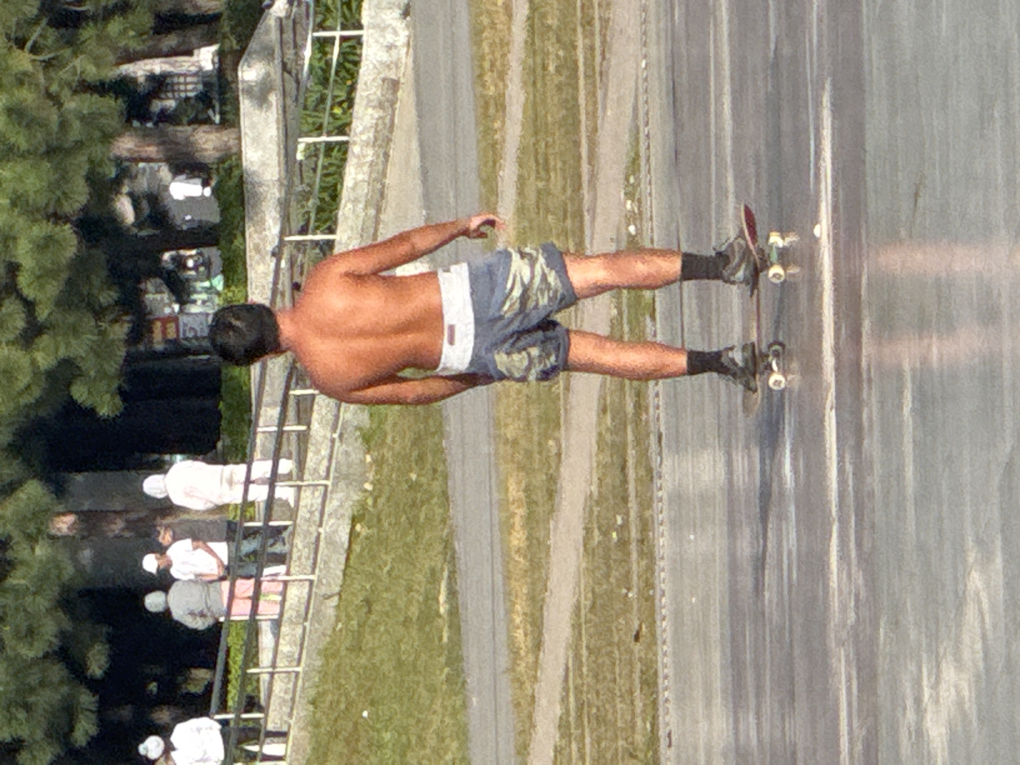 The image shows a young man skateboarding on a wet, paved surface, possibly a park or public square, in Da Lat, Vietnam. He is shirtless and wearing camouflage-patterned shorts with a white waistband, dark socks, and skateboarding shoes. His back is to the camera, and he appears to be mid-movement on his skateboard. In the background, several other individuals are standing near a railing, some of whom are wearing white outfits, suggesting they might be tourists or participants in an event. The environment features lush green trees and a stone wall, indicating an outdoor urban setting. The lighting suggests it is daytime.