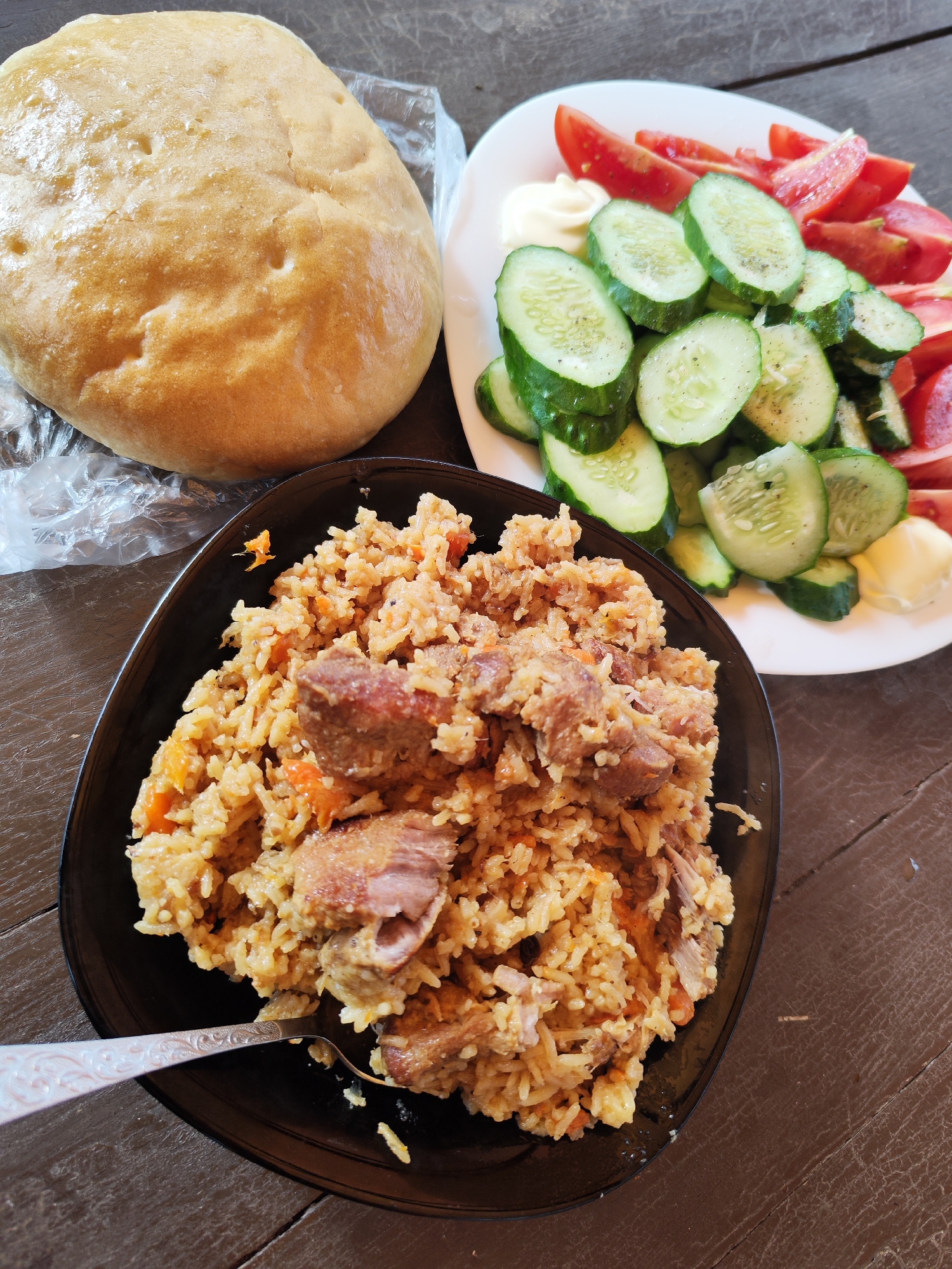 A top-down photographic documentation captures a meal laid out on a dark brown wooden surface, observed in Chaikovsky, Russia.
In the lower-left quadrant of the frame, a black, irregularly shaped bowl holds a substantial portion of pilaf. The pilaf consists of medium-grain rice intermixed with diced orange carrots and irregularly shaped chunks of cooked meat, exhibiting varying degrees of browning and texture. A silver-toned spoon, featuring an embossed handle pattern, is partially submerged within the pilaf, positioned diagonally from the lower-left to the upper-right.
To the upper-right, a white, circular plate presents a fresh salad. This salad comprises numerous thin, circular slices of green cucumber arranged in overlapping layers, and multiple wedge-shaped pieces of red tomato. Two distinct, opaque white semi-solid dollops, consistent with mayonnaise or a dairy-based condiment, are positioned on the plate, one near the tomatoes and one near the cucumbers.
In the upper-left quadrant, a large, spherical loaf of light golden-brown bread, characterized by several surface indentations, rests partially atop a transparent, crinkled plastic bag. The plastic bag extends slightly beneath the pilaf bowl.
The underlying wooden surface displays a dark brown hue, prominent wood grain, and minor surface imperfections consistent with wear and age. No individuals are present in the frame. The scene is illuminated by an overhead light source, casting soft reflections on the bread and the plastic bag.
