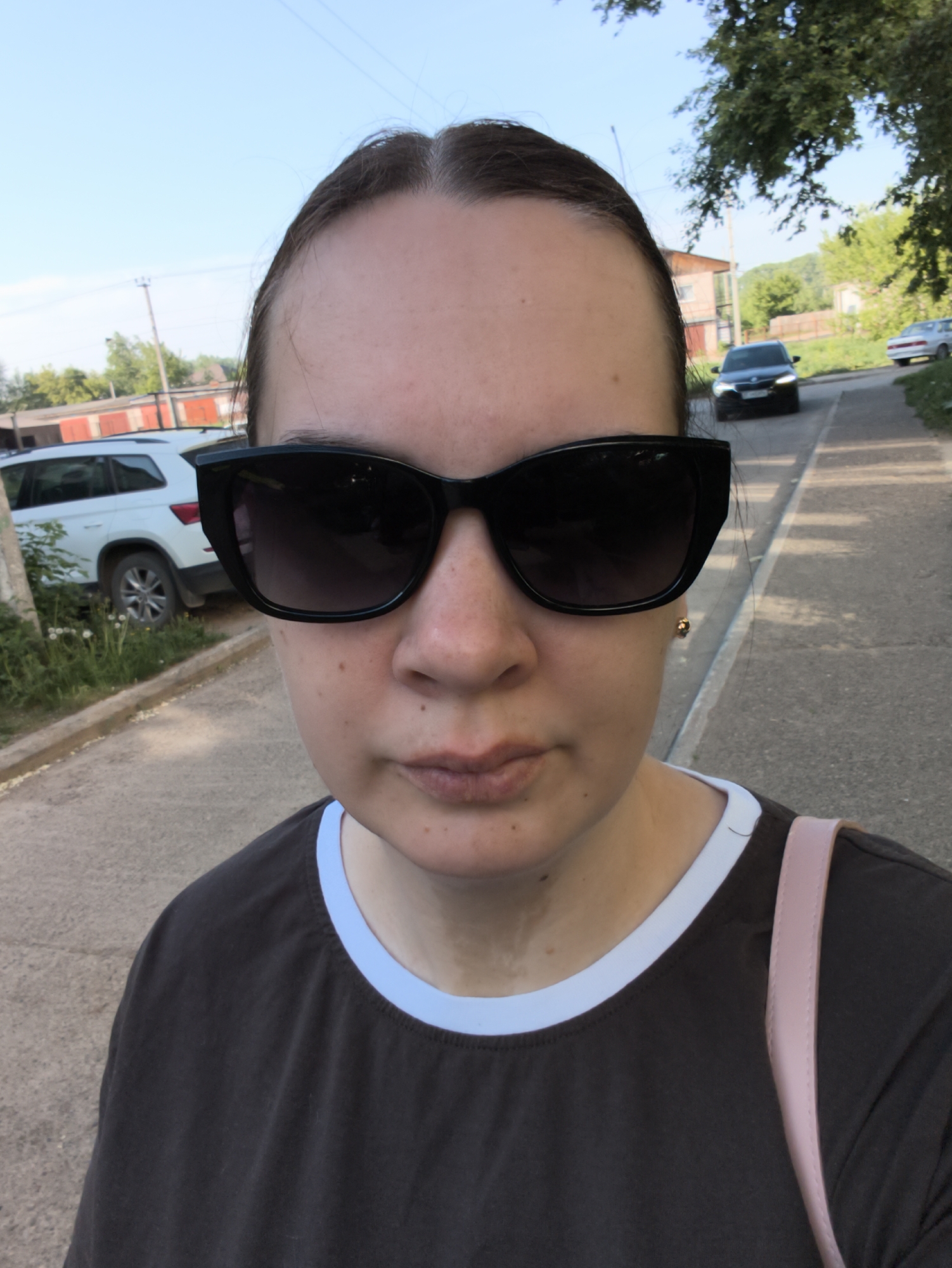 A woman is taking a selfie outdoors on a sunny day. She is wearing dark sunglasses, a brown T-shirt with a light blue collar, and has a pink strap over her shoulder. In the background, there is a street with two cars, one dark and one light, parked on the sides. There is a light-colored building on the left side of the road behind the light car. Trees are visible in the background. The location appears to be in a residential area in the city of Chaikovsky, Russia.