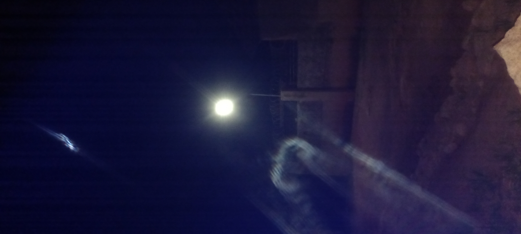 The image is a night-time photograph taken in the city of Kwang, Nigeria. A solitary street lamp illuminates the scene, casting a bright glow. In the sky, a streak of light suggests a passing object, possibly a meteor or a plane. The foreground is dimly lit, with indistinct shapes that could be natural formations or debris. A wall with barbed wire topping runs across the middle ground. The overall atmosphere is dark and mysterious.