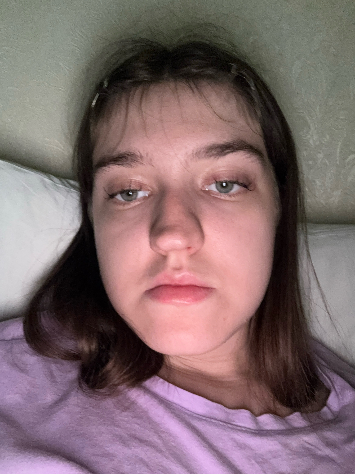 The image shows a close-up, low-angle selfie of a young woman with light brown hair. She is lying down, with her head on a white pillow and her body partially visible, wearing a light purple t-shirt. Her expression appears subdued, with her eyes looking slightly downwards and to the side, and her mouth slightly downturned. The background consists of a textured light green wall. The image was taken indoors, likely in a bedroom. There is no indication of the location being in the city of Zavolzhye, Russia.