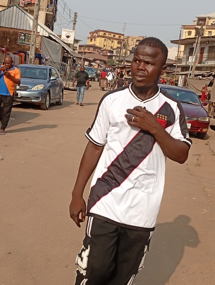 A man in a black and white shirt with black pants stands on a street. The shirt has a large black stripe running from the shoulder to the waist. Buildings and other people are in the background. A blue car and a burgundy car are parked on the sides of the street. The road appears to be unpaved and dusty.