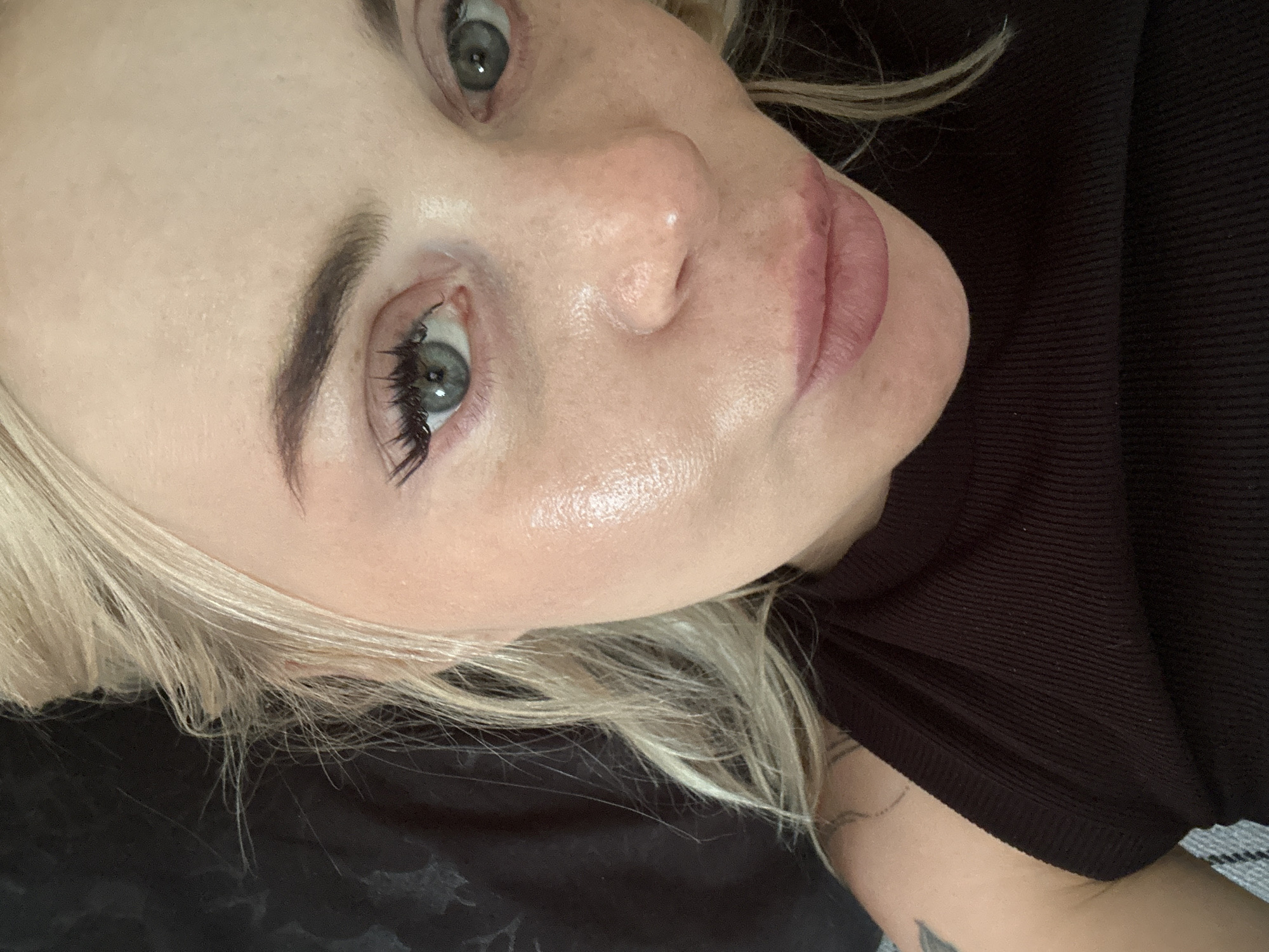 A fair-skinned individual, identifiably female, with blonde hair, is depicted in a close-up, angled portrait, looking directly into the camera. The image focuses on the upper torso, face, and hair. Her blonde hair is loosely styled, with some wispy strands around her face. Her eyes are open, revealing blue-grey irises; the left eye (viewer's right) shows distinct, elongated eyelashes, likely enhanced by mascara or extensions. Both eyebrows are well-defined and dark brown. The skin on her left cheekbone (viewer's right) exhibits a strong reflective sheen, suggesting a dewy complexion or highlighter application. A small, white, raised spot is visible on the tip of her nose. Her lips are full, slightly parted, and tinted a medium pink. She is wearing a dark, ribbed sleeveless top or tank top, possibly black or deep brown. Part of a tattoo, consisting of thin, dark, curvilinear lines, is visible on her upper left arm/shoulder. The background is uniformly dark and out-of-focus, indicating an indoor setting without any identifiable objects or features. The specific city and location are indeterminable from the image.