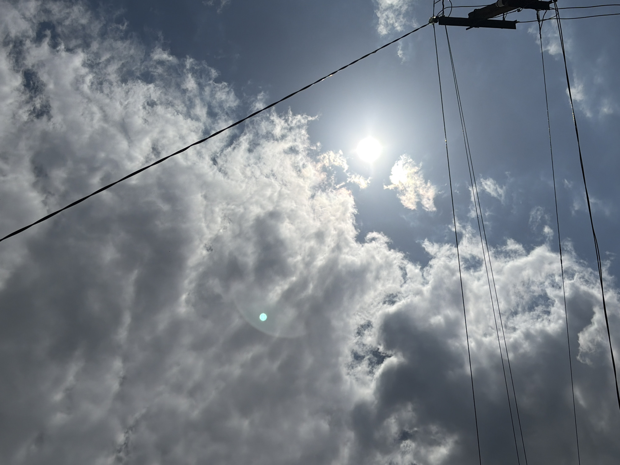 This media file presents an upward view of a bright, partly cloudy sky, characteristic of daytime conditions in Novopokrovskaya, Russia. The sun, a brilliant white orb, is prominently located in the upper central-right portion of the frame, emitting strong light and producing a distinct circular teal-blue lens flare in the lower-left area. The sky features a mix of clear blue patches and scattered clouds; some clouds are bright white and illuminated by the sun, while others appear denser and darker grey.
Several dark, thin wires or cables traverse the image. A thick, dark cable extends diagonally from the upper left corner downwards to the lower right. On the right side, multiple thinner, vertical wires hang from a dark, angular structure, only a portion of which is visible in the top right corner. This structure appears to be overhead infrastructure, possibly part of a utility pole or mast. No people or specific activities are depicted in the scene. The weather is a combination of sunshine and cloud cover. No visible text is present in the image.