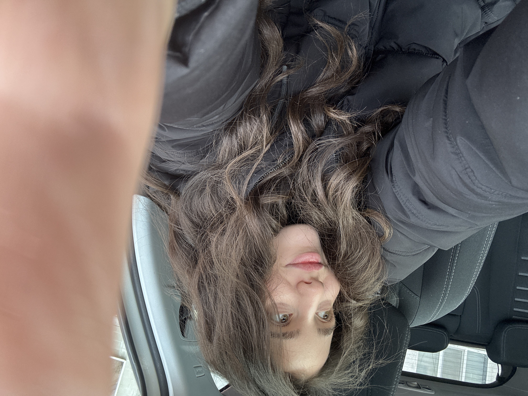 The image shows a woman with long, wavy brown hair wearing a black puffer jacket. She is positioned upside down in the frame, with a blurred object, potentially a finger, visible in the upper left. The background suggests she is inside a car, with parts of the car's interior visible, including a grey dashboard and black seats. The location appears to be urban due to the building visible through the car window. The city location is specified as Unknown, Unknown.