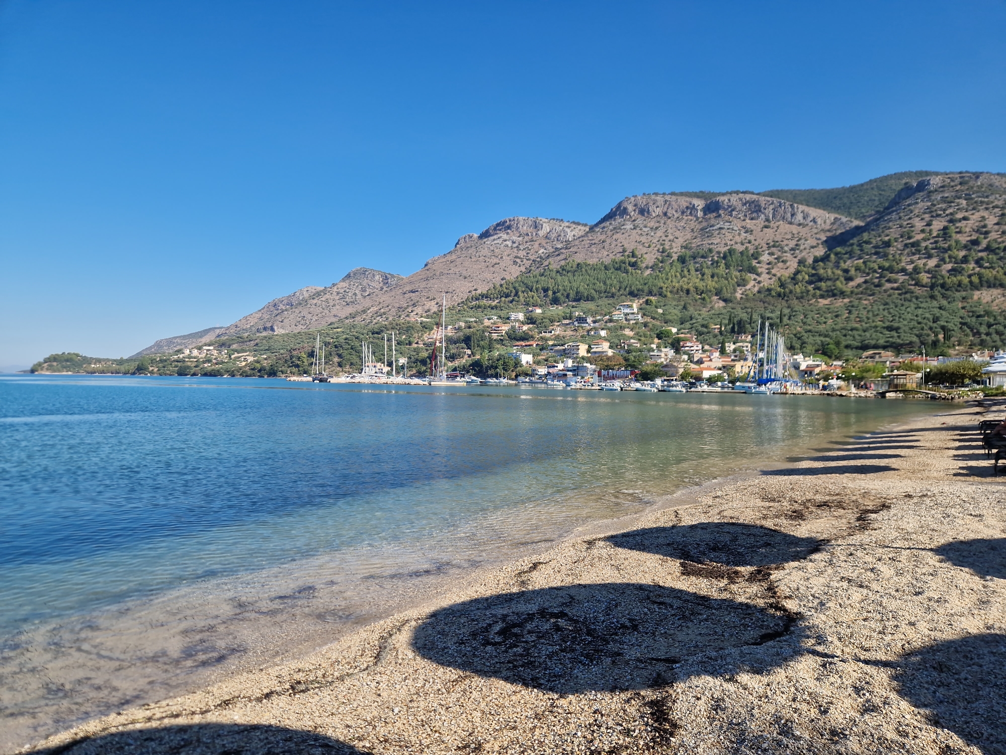 This image depicts a coastal scene in Plataria, Greece, on a clear, sunny day. The foreground is dominated by a sandy and pebbly beach with the gentle lapping of clear blue water. Shadows of beach umbrellas are cast on the sand, suggesting a recreational area. In the middle ground, a calm, blue sea stretches out, dotted with numerous sailboats and yachts moored in a harbor.
The background features a picturesque hillside town built on a slope, with buildings clustered together and interspersed with greenery. The town is backed by a rugged, brown mountain range covered in sparse vegetation, reaching towards the bright blue sky. The overall impression is one of a serene and scenic seaside location, likely a popular tourist destination. There are no visible people in the frame.
