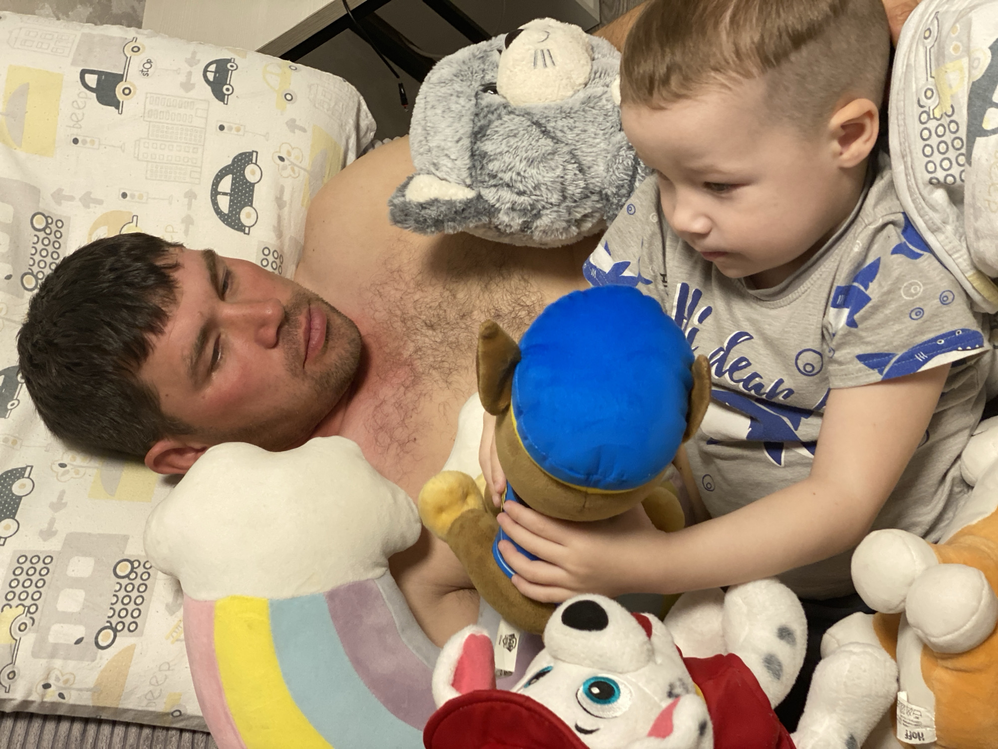 This indoor, eye-level shot captures a domestic scene featuring a man resting on a bed and a young boy playing beside him. The man, with short dark hair and stubble, lies on his back with his eyes closed, suggesting he is sleeping or deeply relaxed. He is shirtless, revealing a hairy chest. He rests on a light-colored pillow patterned with simple drawings of cars (one with "STOP" written on its side), buildings with "00000000" and "000" details, and small text like "beep."
Next to him, a young boy with short, light brown hair sits upright, engrossed in playing with several plush toys. He wears a grey t-shirt adorned with blue shark graphics and partially visible text that might read "Hider." His gaze is directed downwards at the toys in his hands, indicating focus.
The bed is filled with a collection of soft toys. A grey plush toy, resembling a seal or sea lion, rests on the man's chest. The boy holds a brown and blue plush dog, likely Chase from Paw Patrol. Other toys visible include a white and red dog (Marshall from Paw Patrol) and an orange and white dog (Rubble from Paw Patrol), on which a tag showing washing instructions and text "HOFF" is visible. A colorful rainbow-striped cloud plush toy is also seen near the man's side.
The overall atmosphere is calm and intimate, suggesting a moment of rest and playful interaction between the two individuals. The scene is set in what appears to be a bedroom in Sochi, Russia.