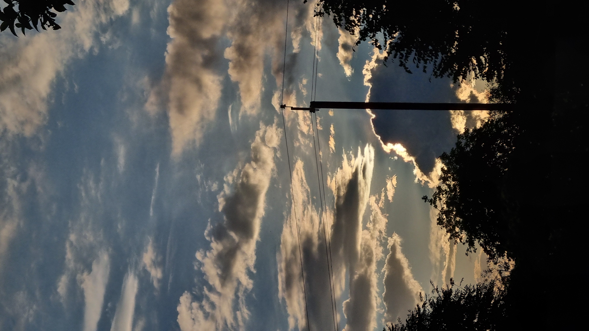 A vertical shot captures a dramatic sky over Vysokoe, Russia, at dusk. Silhouetted trees form a dark border along the bottom of the frame, with a single utility pole standing vertically in the mid-ground, bisecting the scene. Overhead, a complex cloudscape dominates, with streaks of sunlight piercing through gaps in the dark, heavy clouds. The sun's rays illuminate the edges of the clouds, creating a contrast between light and shadow that suggests the sun is low on the horizon. Two parallel wires stretch horizontally across the frame, strung between unseen points, adding a man-made element to the natural scene. The overall impression is one of a dynamic and slightly ominous twilight.