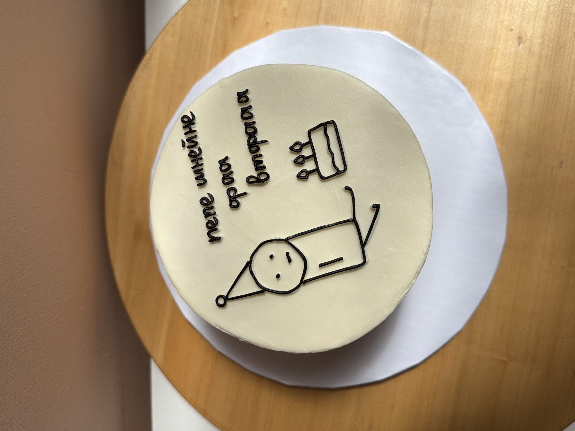 A light cream-colored, round cake with black icing decorations is centered on a white circular cake board. The cake board rests on a light-colored wooden circular surface, likely a cake turntable or table. The cake's top features a simple stick-figure drawing of a person wearing a party hat on the left, depicted with a straight line mouth and two dots for eyes. To the right of the stick figure, a small, two-tiered cake with three candles is drawn. Above the small cake, black icing spells out non-English text across two lines, reading "тепе шинейне" on the upper line and "фраа вторая" on the lower line. The background consists of a plain, light brown wall.