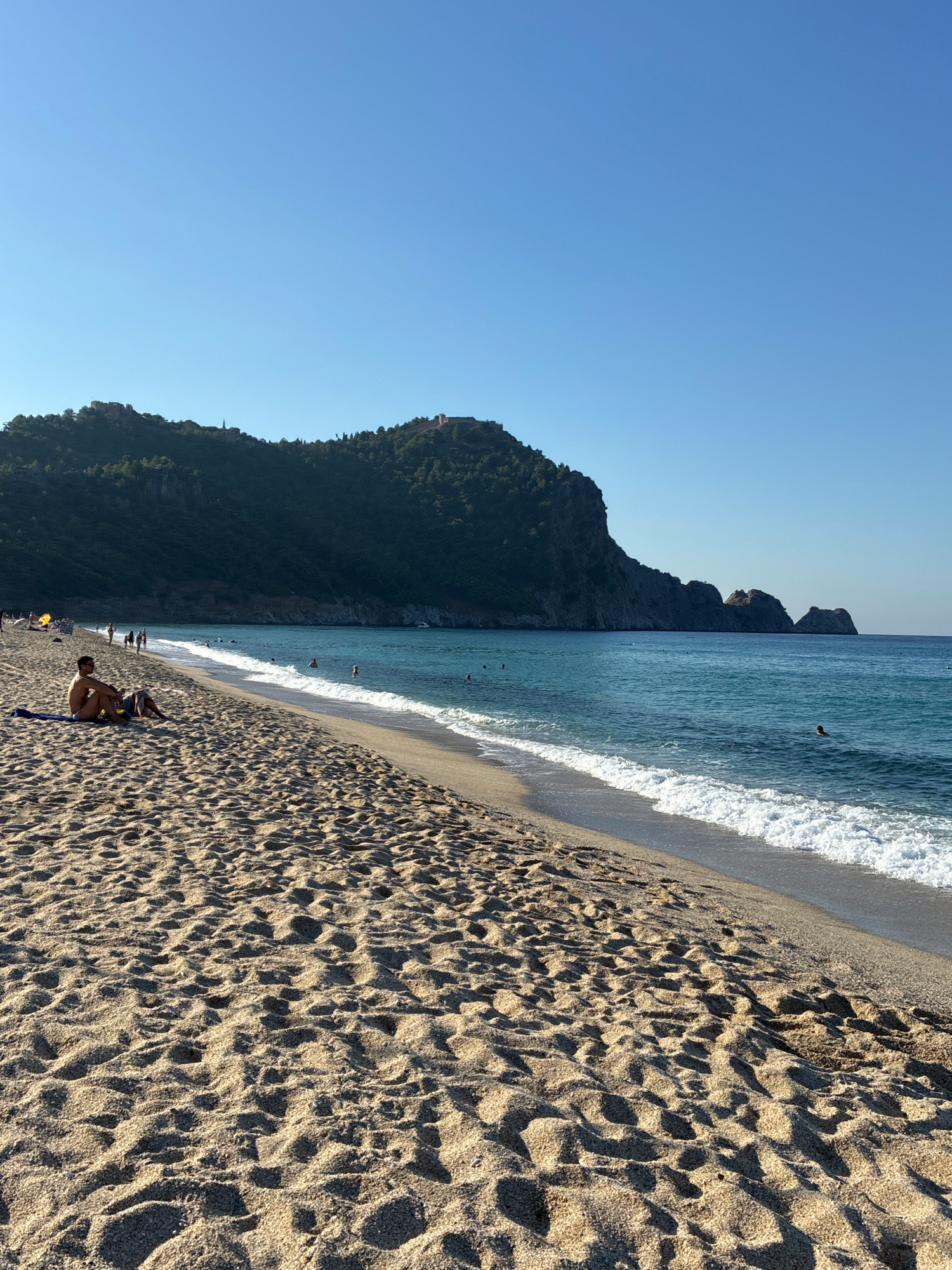 The image depicts a sandy beach on a sunny day, with the turquoise Mediterranean Sea in the background. The beach is populated by a few individuals, some of whom are sunbathing on towels, while others are in the water. The sea exhibits moderate waves with white foam. In the distance, a lush green hill rises, crowned with the ancient Alanya Castle, a prominent landmark of the city. The sky is a clear, cloudless blue, indicating fair weather conditions.