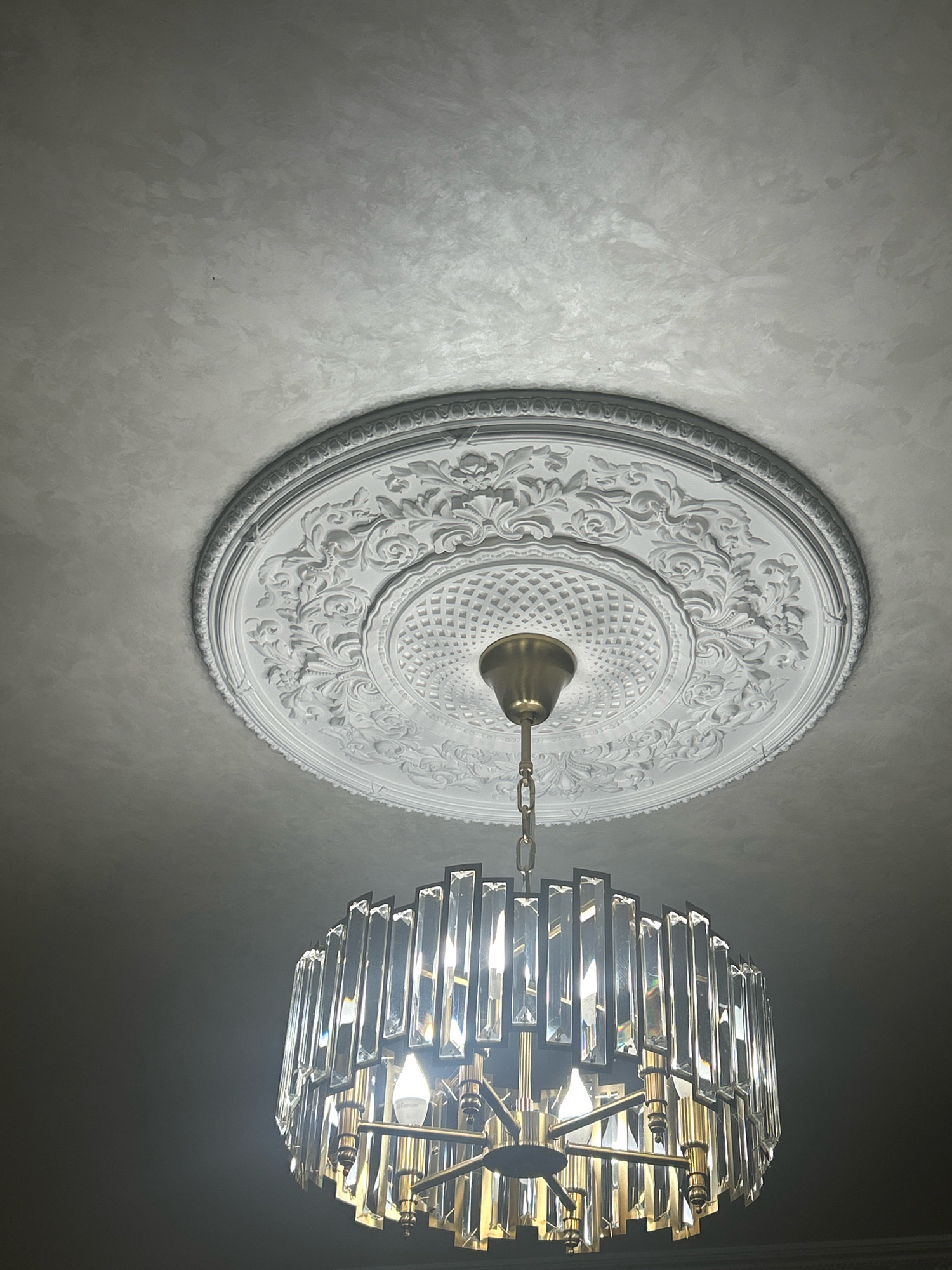 The image displays a chandelier hanging from a ceiling. The ceiling features ornate white plasterwork, including a decorative medallion. The chandelier itself is modern, with a brass-colored frame and numerous clear glass or crystal elements that refract light. The glass elements are rectangular and arranged vertically around the frame, creating a sparkling effect. The bulbs within the chandelier are illuminated. The ceiling appears to be painted in a light, textured finish. The scene is indoors, and no people or specific exterior environmental elements are visible. The location of the image is identified as Grozny, Russia.