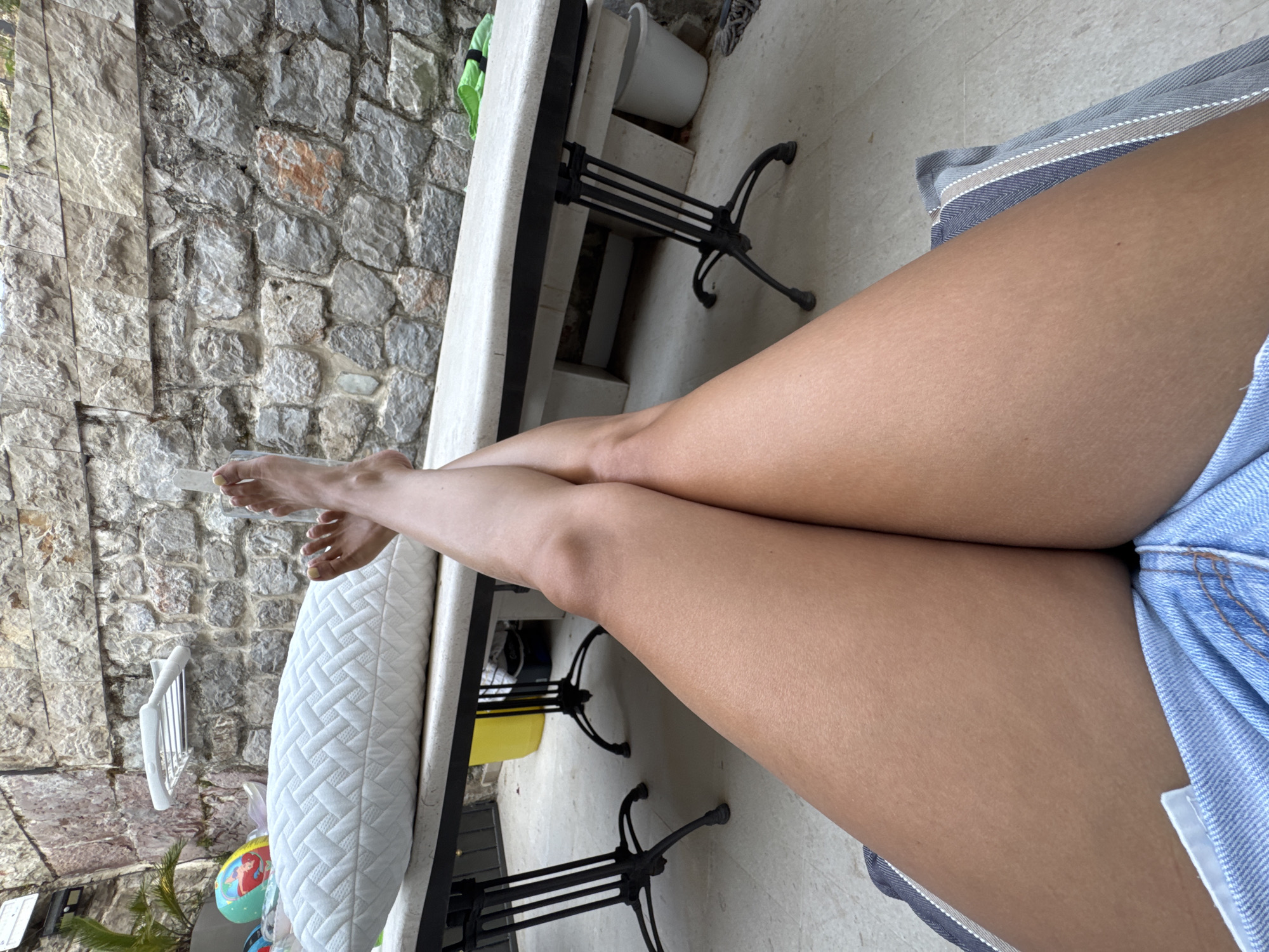 A low-angle, close-up shot shows a person's tanned legs crossed at the thighs, resting on a striped cushion. The person is wearing light blue denim shorts that are mostly obscured by their legs. The legs are smooth and appear to be sun-kissed. Behind the legs, a white quilted pillow sits on top of a long, narrow table with a dark metal base. The table is positioned outdoors, in what appears to be a patio or courtyard area. The background consists of a rough, stone wall, typical of older architecture. To the left of the table, a palm frond can be seen, and further left, a colorful inflatable ball with an image of Ariel from "The Little Mermaid" is partially visible. The ground is a light-colored, textured surface.