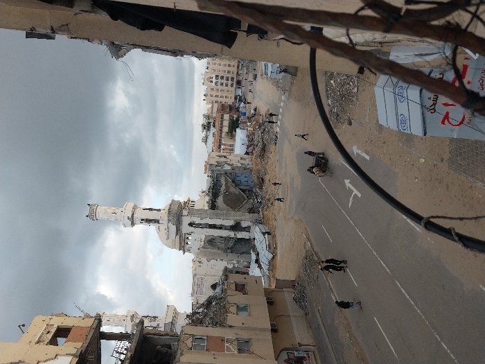 The image depicts a street scene in Khan Yunis, Palestinian Territory, under an overcast sky. The foreground is partially obscured by dark wires and a brown metal frame. To the immediate right, a heavily damaged building shows exposed rebar and broken concrete, with dark fabric hanging from an upper floor. Below this, a large white sign with red and black Arabic text is visible, partially obscured.
The central feature is a paved road, marked with white dashed and solid lines, and two white turn arrows indicating forward and right. The road is flanked by sandy, debris-strewn ground. In the middle distance, a white donkey pulls a two-wheeled cart, with a person walking alongside it on the right. Further to the left on the road, three men are walking together. Scattered figures, including adults and possibly children, are visible on the sandy areas and further down the street.
In the background, a severely damaged mosque with a prominent white minaret stands partially intact, surrounded by piles of rubble and structural remnants. Its dome and upper sections show significant damage. To the left of the mosque, a multi-story building is extensively damaged, with large sections of its facade missing, revealing interior spaces and structural components. Similar damaged buildings line both sides of the street further into the distance, contributing to an overall appearance of widespread structural destruction. The street itself appears to be a thoroughfare, with more figures visible walking along it in the distance, amidst the ruins.