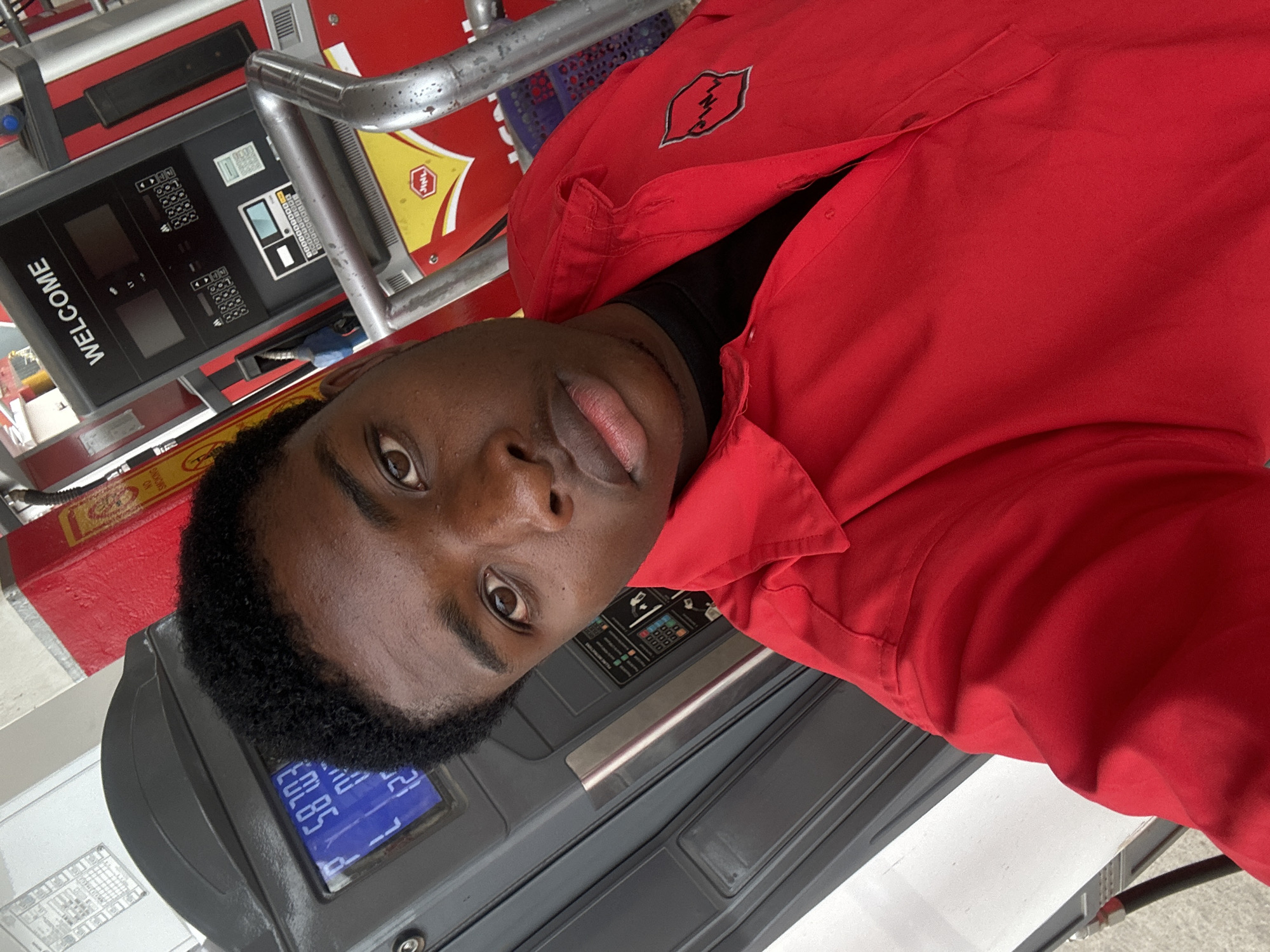 A young man is taking a selfie at a gas station. He is wearing a red shirt with a logo on the left breast. Behind him is a gas pump showing a fuel amount of 58.85. The pump also has a "WELCOME" message displayed. Based on other visual cues, the location is likely in Jos, Nigeria.