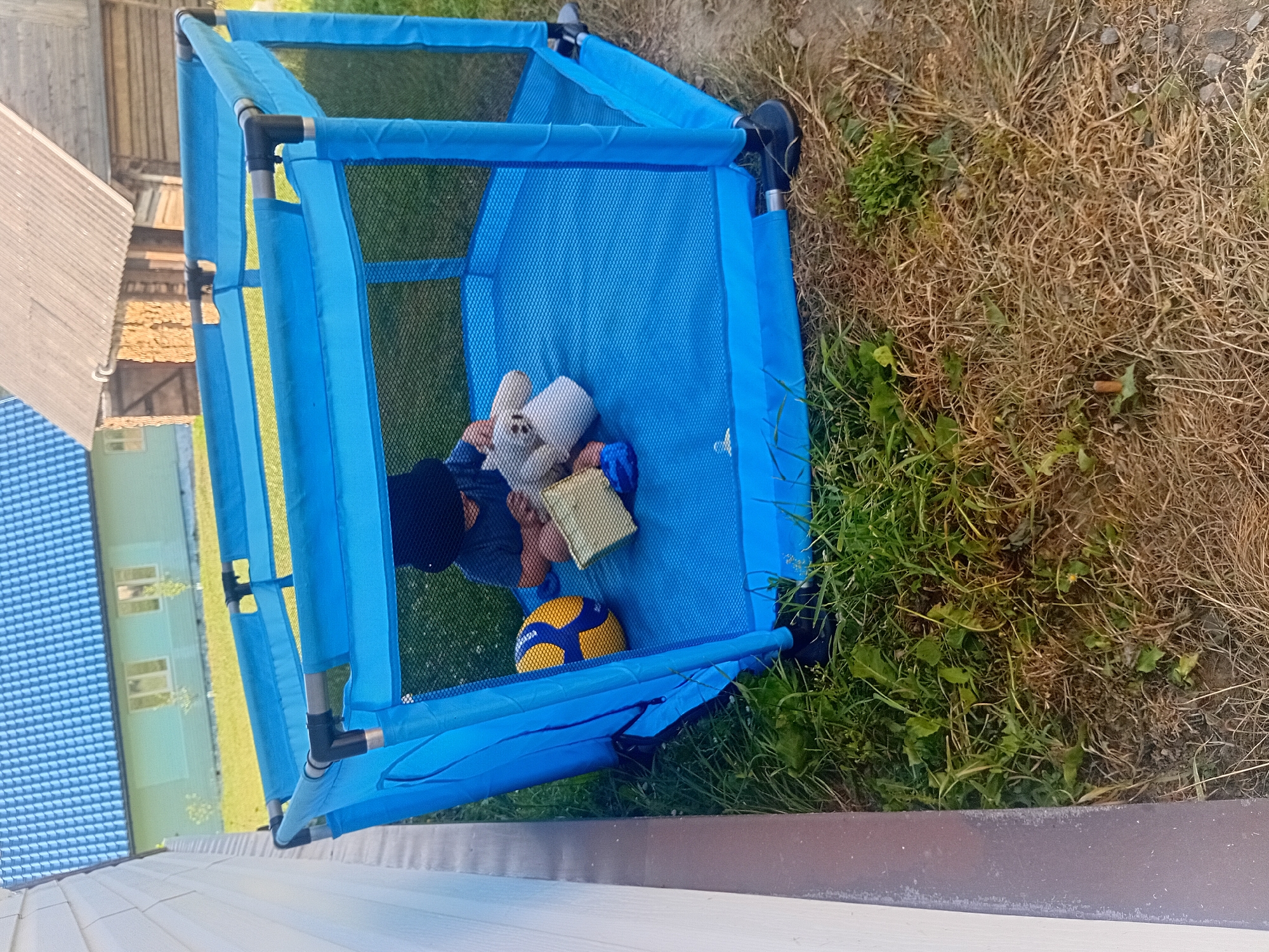 A child wearing a dark hat and shirt sits inside a blue playpen filled with toys. The playpen is located outdoors on a patch of grass, with dry, brown vegetation in the foreground. In the background, a light green house with a blue roof is visible, along with a wooden structure. The scene appears to be in a rural or suburban area, possibly Verkhniachka, Ukraine, given the architectural style and vegetation.