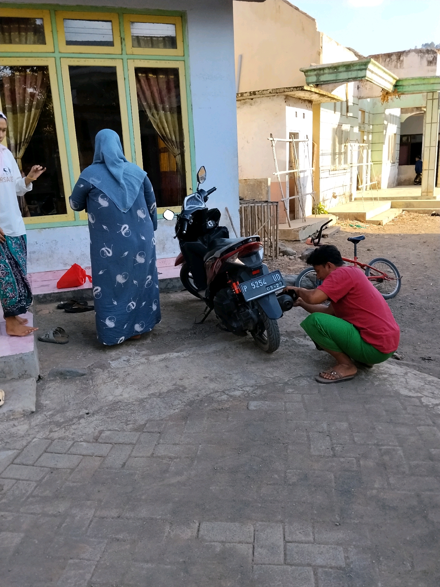 In Panggungan, Indonesia, a man wearing a red t-shirt and green pants is squatting next to a dark-colored scooter, appearing to work on its exhaust pipe. He is wearing sandals. A red bicycle with a bent frame is parked behind him. To the left, two women are standing near a building with yellow-trimmed windows. One woman, wearing a blue hijab and a patterned dress, is facing away from the camera. The other woman, partially visible, is wearing a white top and patterned pants. A red bag is on the ground near the women. The ground is a mixture of concrete and paved bricks. The environment appears to be outdoors, possibly a residential area, with partially constructed buildings visible in the background. The sky is clear and sunny.