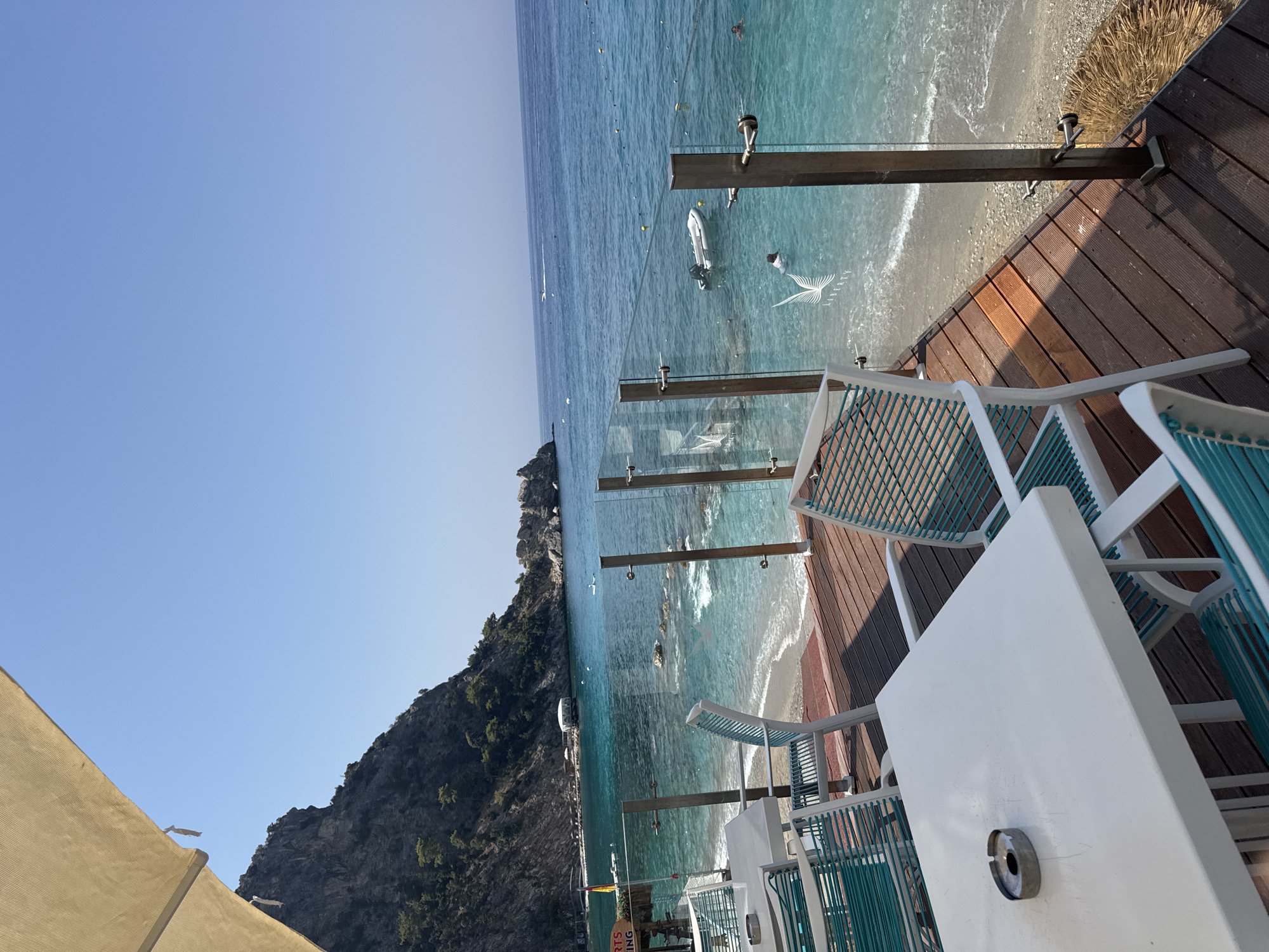 This is a photograph of a beachfront restaurant or cafe in Fethiye, Turkey. In the foreground, there is a white rectangular table with an ashtray on it, surrounded by chairs with white frames and teal webbing. The seating area is on a wooden deck overlooking a beach. The beach has fine pebbles and is bordered by clear turquoise water.
Beyond the deck, the sea stretches out under a clear blue sky. Several boats, including a small inflatable one and a speed boat in the distance, are visible on the water. A few people are swimming or wading in the sea. In the middle ground, there is a rocky coastline with some vegetation. A steep, tree-covered mountain dominates the left side of the image. The overall impression is a sunny, tropical vacation setting.