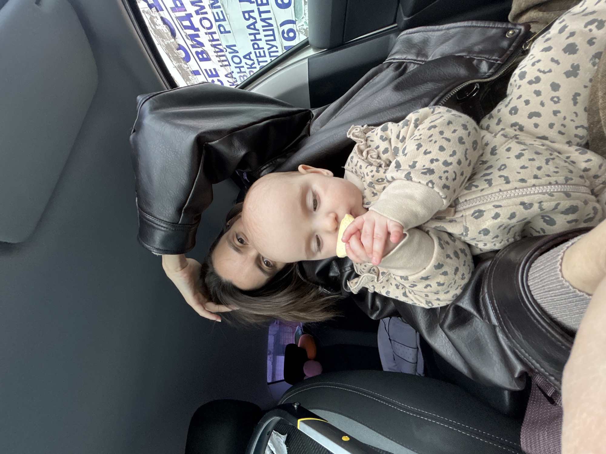 A baby in a leopard-print onesie is being held by a woman in a car. The woman is wearing a brown leather jacket. The baby is holding a piece of food in its hand. The car is parked in Moscow, Russia. A sign in Russian is visible in the background.