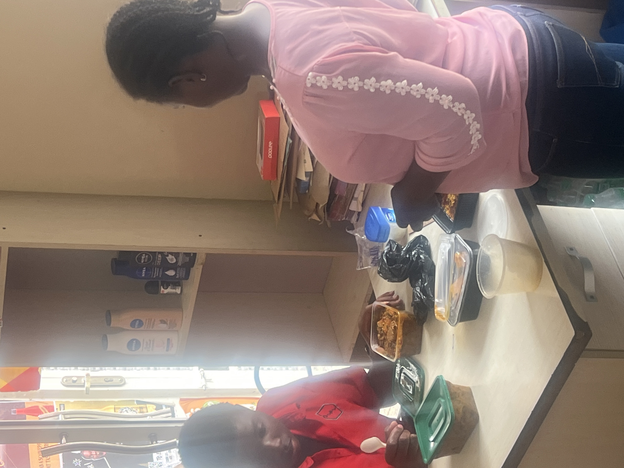 The image, rotated clockwise, depicts an indoor scene with two individuals interacting with food containers on a table or counter.
On the left, a person wearing a red t-shirt is shown, holding a white plastic spoon over a green food container, appearing to be eating. Their head is angled downwards, focusing on the contents.
On the right, a second person, dressed in a pink long-sleeved top adorned with white floral embellishments on the sleeves and dark blue jeans, stands looking towards the table. On the table, several food containers are visible: a clear plastic container with a stew-like dish, a black plastic container, another green container (partially obscured), and a translucent cup. A black plastic bag and a blue plastic bottle are also present.
In the background, a shelf unit holds various items. On the upper shelf, multiple bottles of Nivea lotions (in dark blue, beige, and white colors) and a Nivea Men deodorant stick are arranged. Below this, a red box with the text "COOLERE" (or similar) is placed on top of some papers. Further down, a window or opening with a metal latch is visible, along with some posters or documents on the wall.
Both individuals have dark skin tones. The scene suggests a casual mealtime in a functional space, possibly a small eatery, break room, or shop, consistent with a setting in Jos, Nigeria. The time of day is not explicitly indicated but appears to be well-lit indoors.
