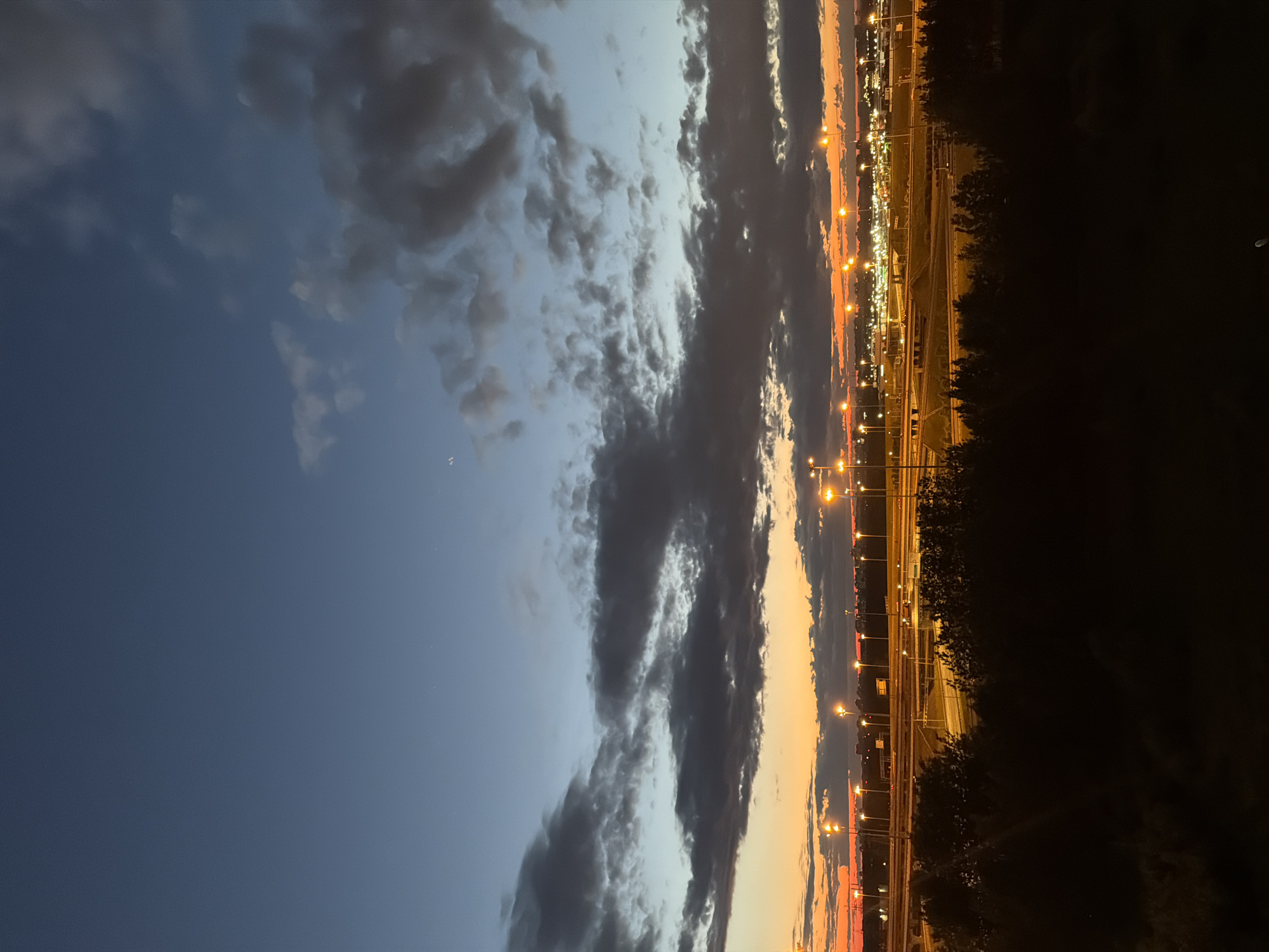 This is a photograph taken at dusk, with a twilight sky transitioning from deep blue at the top to orange and red near the horizon. Dark, scattered clouds are visible in the sky. Below the sky, a landscape unfolds, featuring a silhouette of trees in the foreground, obscuring much of the ground. Beyond the trees, an urban or industrial area is illuminated by numerous streetlights, casting a warm glow. This area appears to be a complex network of roads, possibly a highway interchange, with buildings and structures visible in the distance. The overall scene depicts an evening view of a city or developed area, with the last light of day meeting artificial illumination.