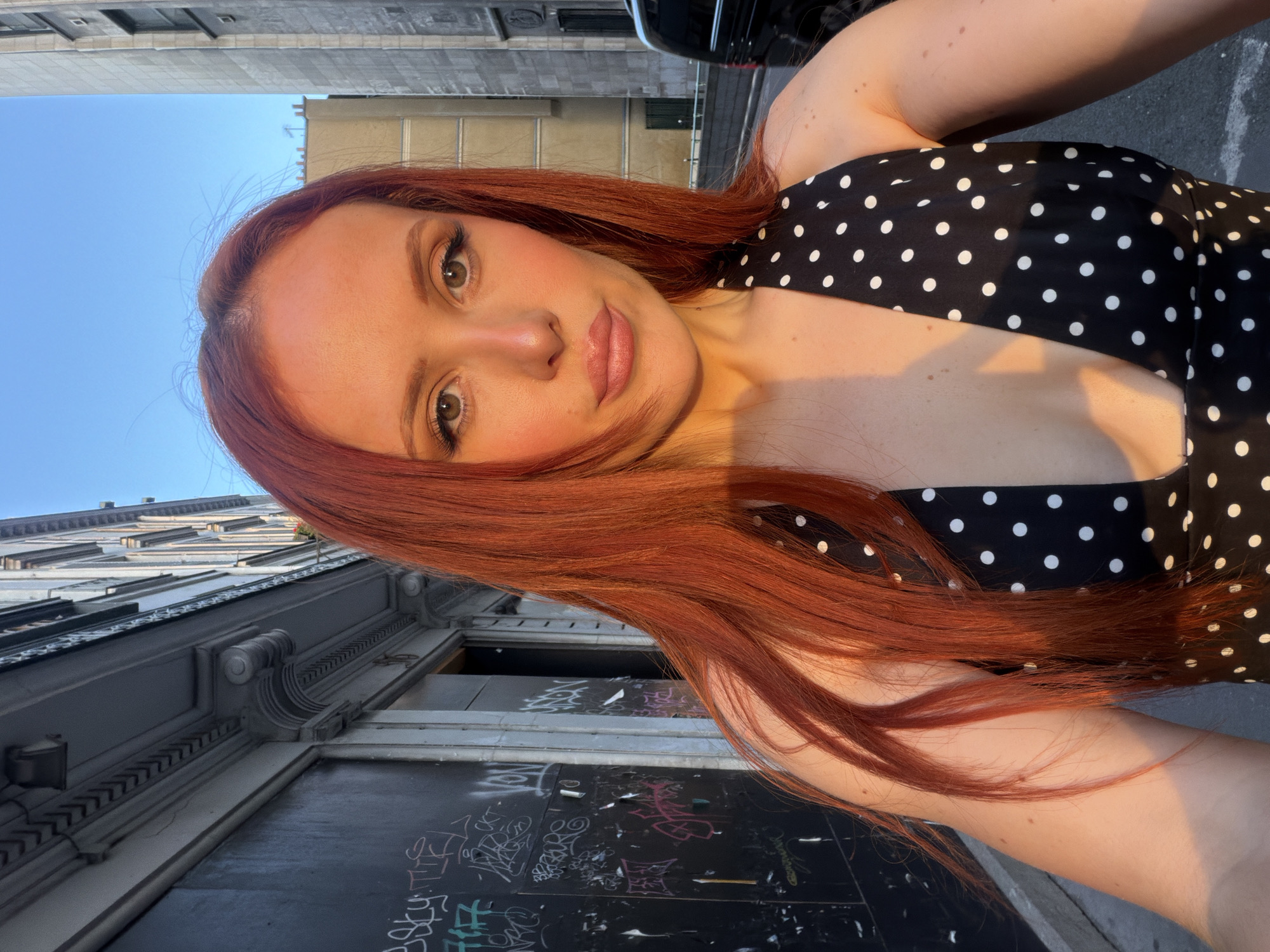 The image shows a selfie of a young woman with long red hair, wearing a black and white polka dot dress. She is standing on a street in Paris, France. Behind her are buildings with ornate architectural details, including cornices and decorated windows. The lower portion of the building nearest to her has graffiti on the wall. The sky is blue. The lighting suggests it is either early morning or late afternoon.