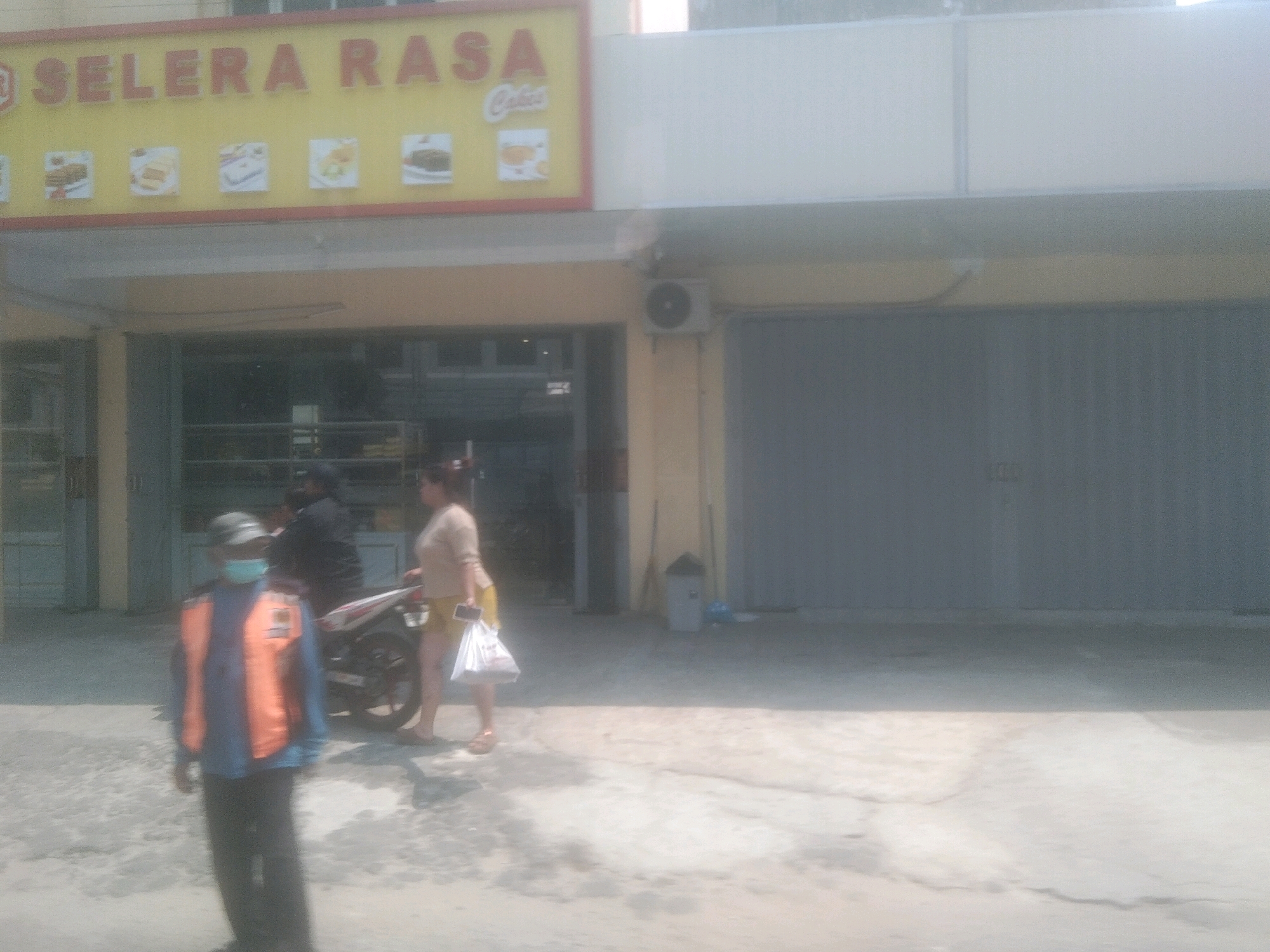 The image depicts the exterior of a shop with a prominent yellow sign that reads "SELERA RASA CAKES". Several small framed pictures of cakes are displayed on the sign. In front of the shop, a person wearing a blue shirt, an orange vest, and a face mask stands in the foreground, facing the camera. Behind them, another person in a black helmet and jacket is partially visible, along with a woman in a beige top and yellow shorts holding a shopping bag and a phone. A motorcycle is parked nearby. The ground is paved with concrete, and the building has large metal roll-up doors on the right side. The scene appears to be set in a commercial area in Bandar Lampung, Indonesia.