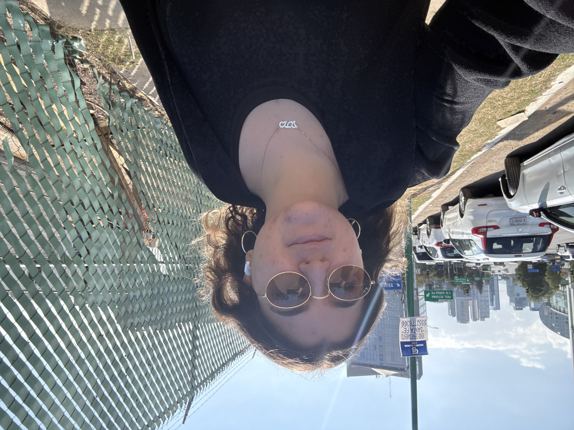 An outdoor, eye-level selfie-style photograph captures a young female subject on a sunny day in Mexico City's Santa Fe district. The subject, positioned centrally, has shoulder-length wavy brown hair with lighter highlights and wears round, gold-rimmed sunglasses, white wireless earbuds in both ears, silver hoop earrings, and a silver necklace with the Hebrew word "מאיא" (Maya) scripted on it. She is dressed in a black top and a black jacket.
To the subject's immediate right, a green chain-link fence with woven privacy slats extends from the foreground into the mid-ground. To her left, a paved sidewalk transitions to a dirt and grass verge, followed by a multi-lane road. Numerous vehicles, predominantly white, are visible on the road, some parked alongside the verge, including a white SUV/minivan and a silver sedan, and others in motion further down the road.
The background features a modern urban skyline dominated by multiple high-rise buildings and skyscrapers. A tall green pole on the left displays a blue sign advertising a Coldwell Banker apartment for sale in "SANTA FE" with the phone number "5552778366," and below it, a green directional road sign pointing to "PROL. REFORMA Y DE GUADAL PJE." The Hilton Santa Fe hotel building is also identifiable within the background architecture. The sky is clear blue with scattered white clouds, and bright sunlight casts distinct shadows.