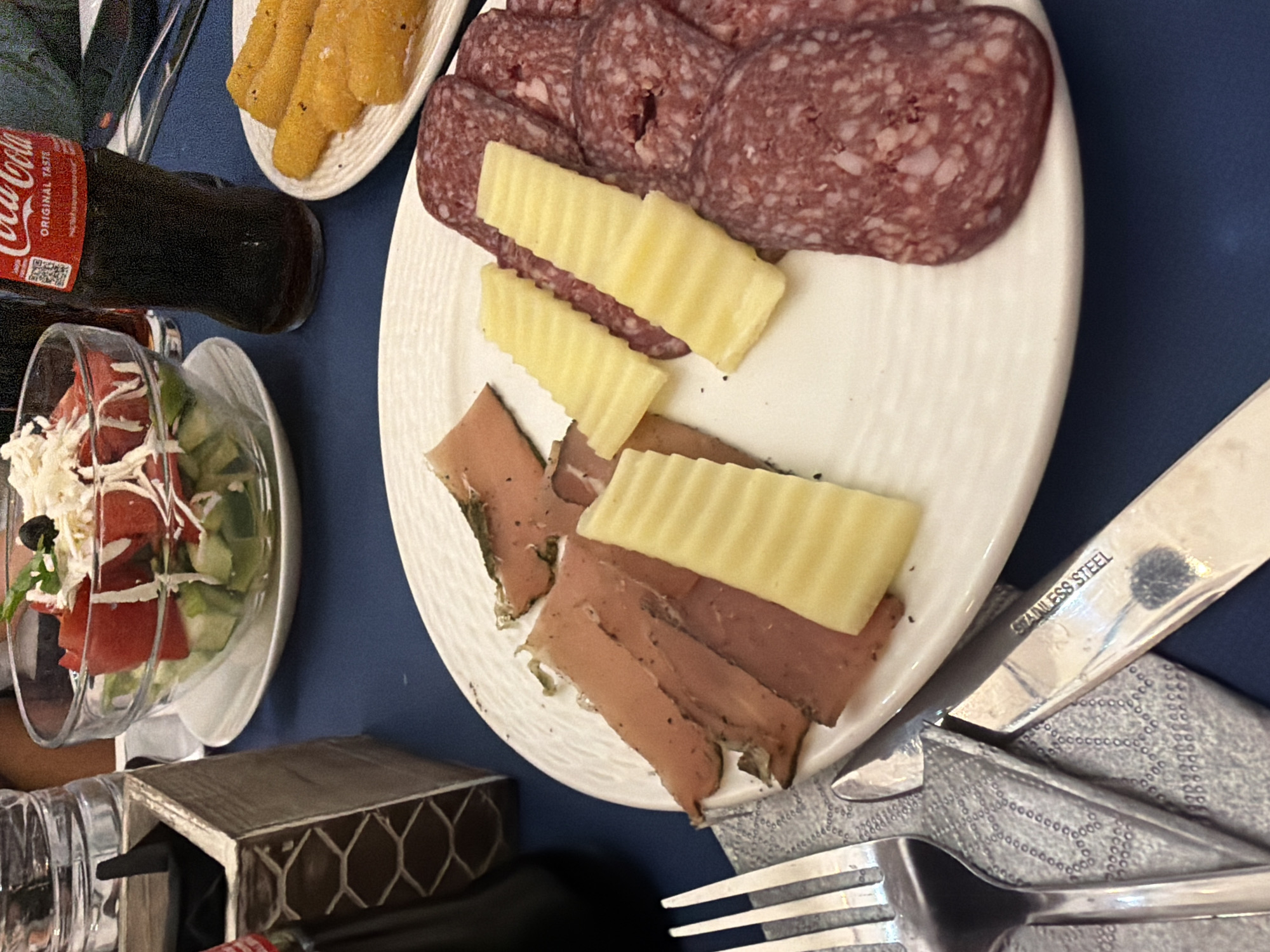 The image depicts a table setting at what appears to be a restaurant. On the table, a white plate holds an assortment of food including thinly sliced cured meats, cheese slices with a ribbed texture, and a small salad in a glass bowl containing chopped tomatoes, cucumbers, and shredded white cheese, topped with a black olive and some greens. To the right of the salad bowl, a bottle of Coca-Cola with its iconic red label is visible. In the foreground, cutlery, including a fork and a knife, are placed on a blue tablecloth next to a patterned napkin. A textured container with dark contents is positioned to the left of the salad bowl. The lighting suggests an indoor setting, possibly during the day or evening. The overall scene suggests a meal is being served. The city of Roman, Bulgaria, is not depicted in this image.