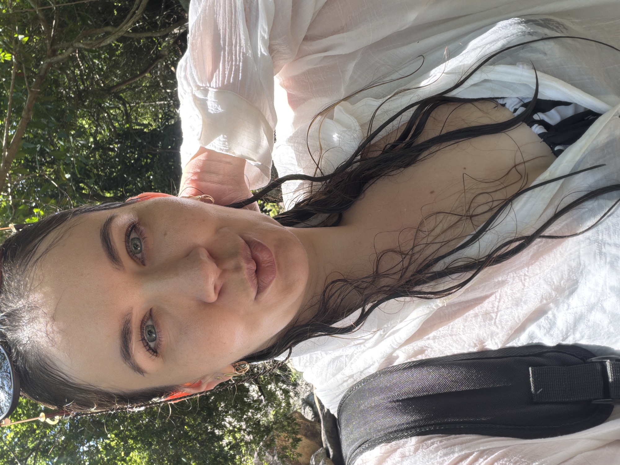 A woman with wet, dark hair, wearing a white shirt and a black and white patterned bikini top, takes a selfie outdoors. She is holding her hand to her neck and her hair is falling around her face. A black backpack is visible over her left shoulder. She is outdoors in a natural setting with lush green trees and foliage in the background, suggesting a location near water or a natural park. This is likely Hoa Thang, Vietnam, given the vegetation and natural environment.
