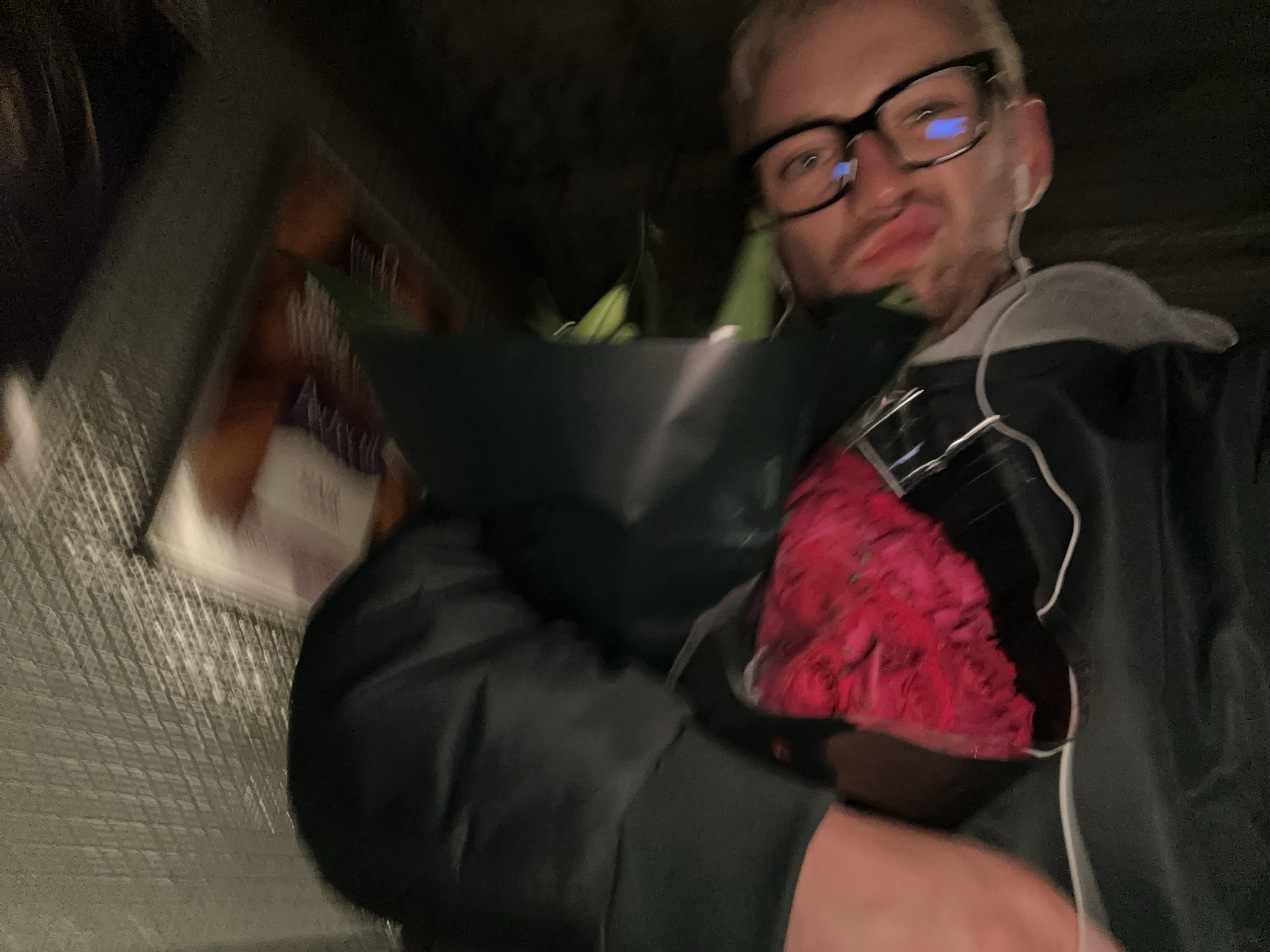 A person with fair skin, blonde hair, and wearing black-rimmed glasses is holding a bouquet of red flowers. They are also wearing wired earphones and a dark grey hoodie with the hood down. The scene appears to be dimly lit, and the background is blurry, with a corrugated-looking surface visible to the left. The person is making a kissing face expression. The location is not identifiable as London, United Kingdom, based on the visual information provided.