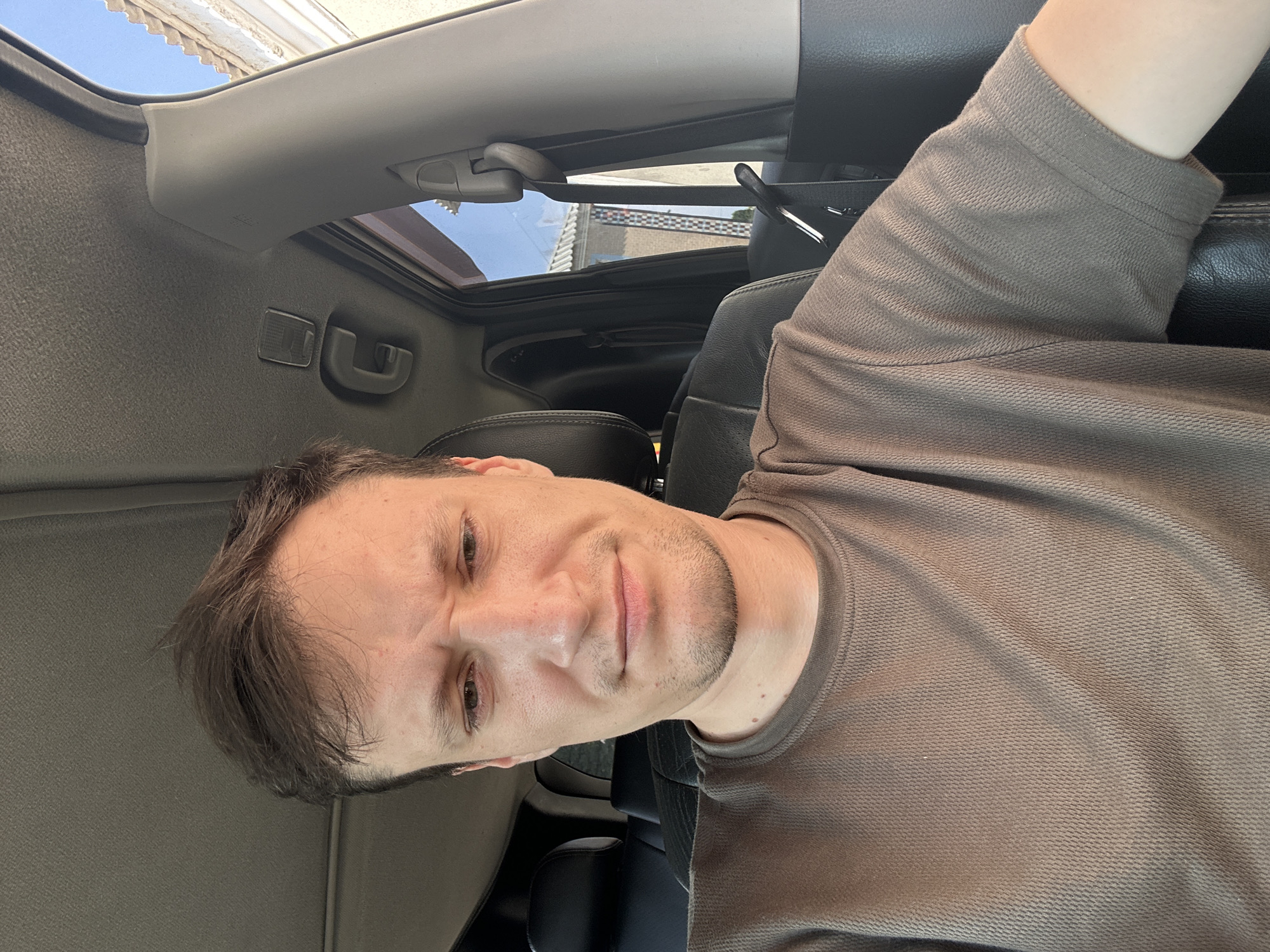 A man is seated in the driver's seat of a car, looking forward at the camera. He has short brown hair and a light beard. He is wearing a brown t-shirt. The interior of the car is visible, including the dashboard, ceiling, and seats. The background shows buildings and a blue sky, suggesting an outdoor urban environment. The image is taken from a slightly elevated, front-facing angle. There is no indication that the image was taken in Shatava, Ukraine.