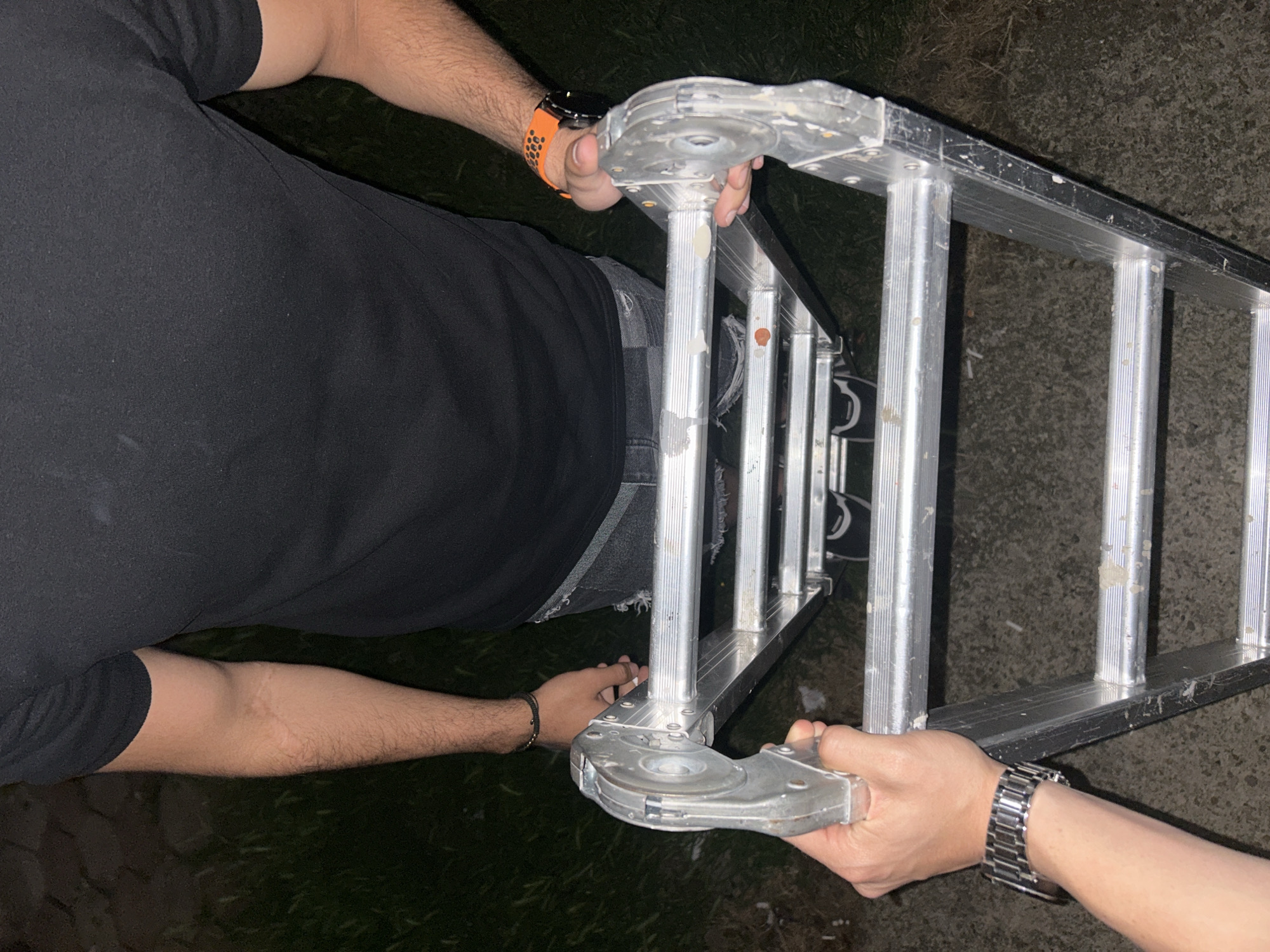A person is holding an aluminum ladder horizontally in an outdoor setting, possibly a backyard or driveway. The person is wearing a black t-shirt and grey jeans. The person's left hand is visible with an orange watch. Another person wearing a silver watch holds the other end of the ladder. The environment around them appears to be dimly lit, with what looks like grass. The city of Elhovo, Bulgaria is not identifiable from the image.