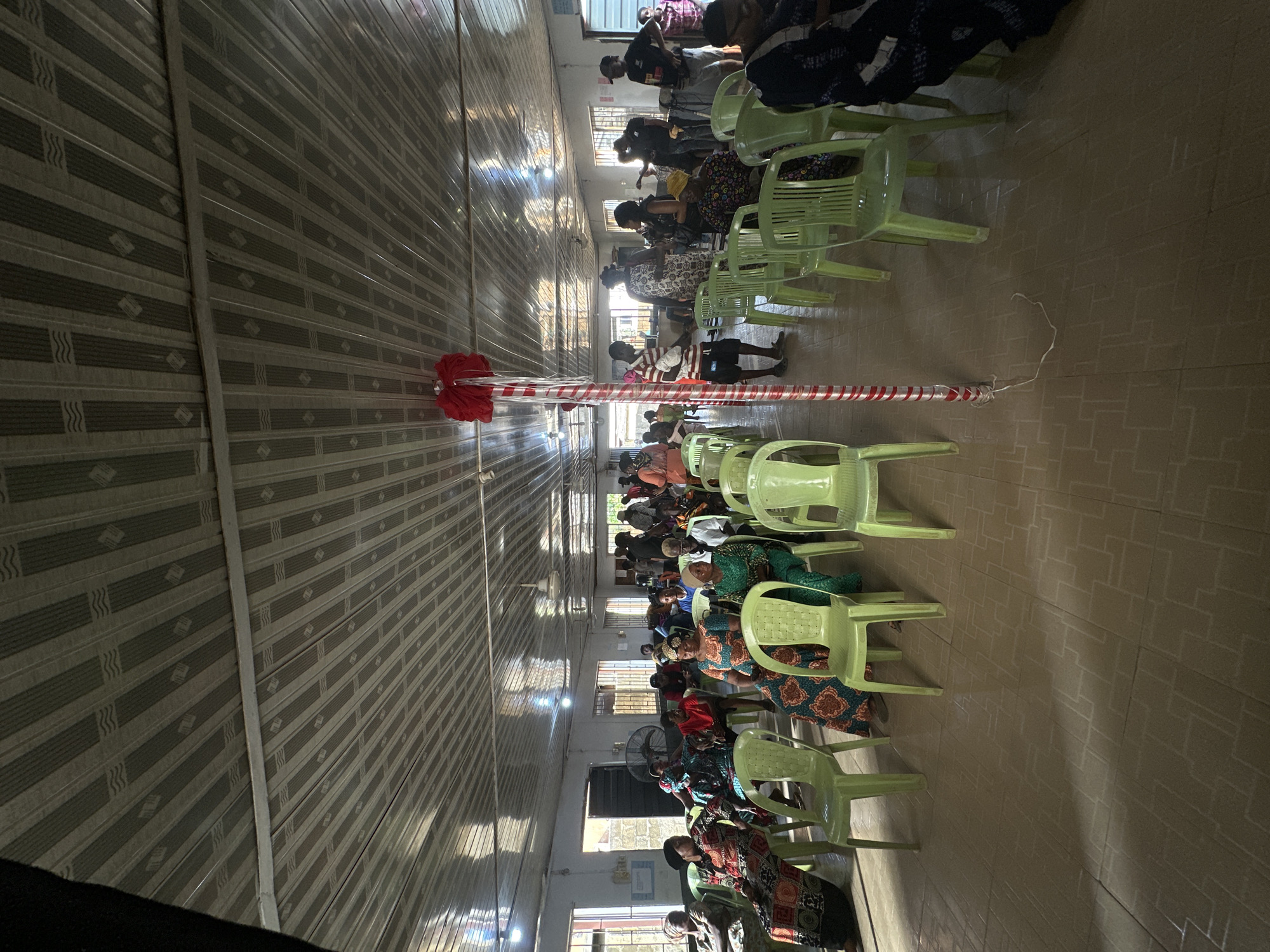 The image shows a large interior space, likely a church or community hall in Lagos, Nigeria. Rows of light green plastic chairs are arranged facing the center of the room, where a pole wrapped in red and white striped tape stands. People are seated in the chairs, wearing a variety of clothing patterns and styles. Windows are visible along the walls, providing natural light. The ceiling is covered with patterned metal panels.