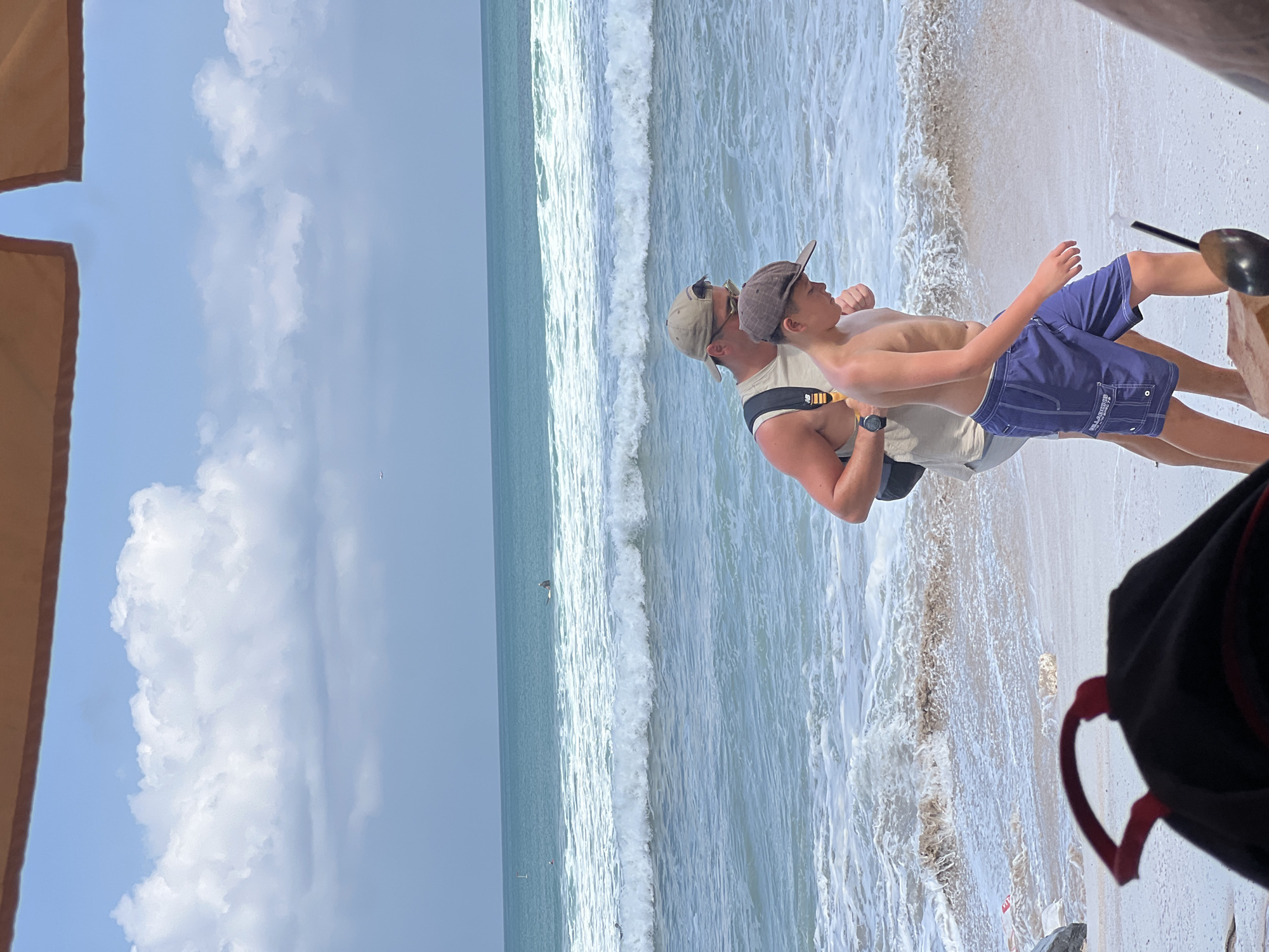 A man and a boy walk on a wet, sandy beach in Kuta, Indonesia, with gentle waves and white foam lapping at the shore. The ocean stretches out to the horizon under a hazy sky with scattered white clouds. Both individuals are facing right, with the man in a light-colored tank top and a tan baseball cap walking behind and slightly to the left of the boy, who wears blue swim trunks and a brown baseball cap. The man has a backpack on, visible with black straps and a yellow accent. In the background, a few surfers are visible in the ocean. In the foreground, part of a black backpack with red straps is visible, along with a brown object that may be a piece of wood.